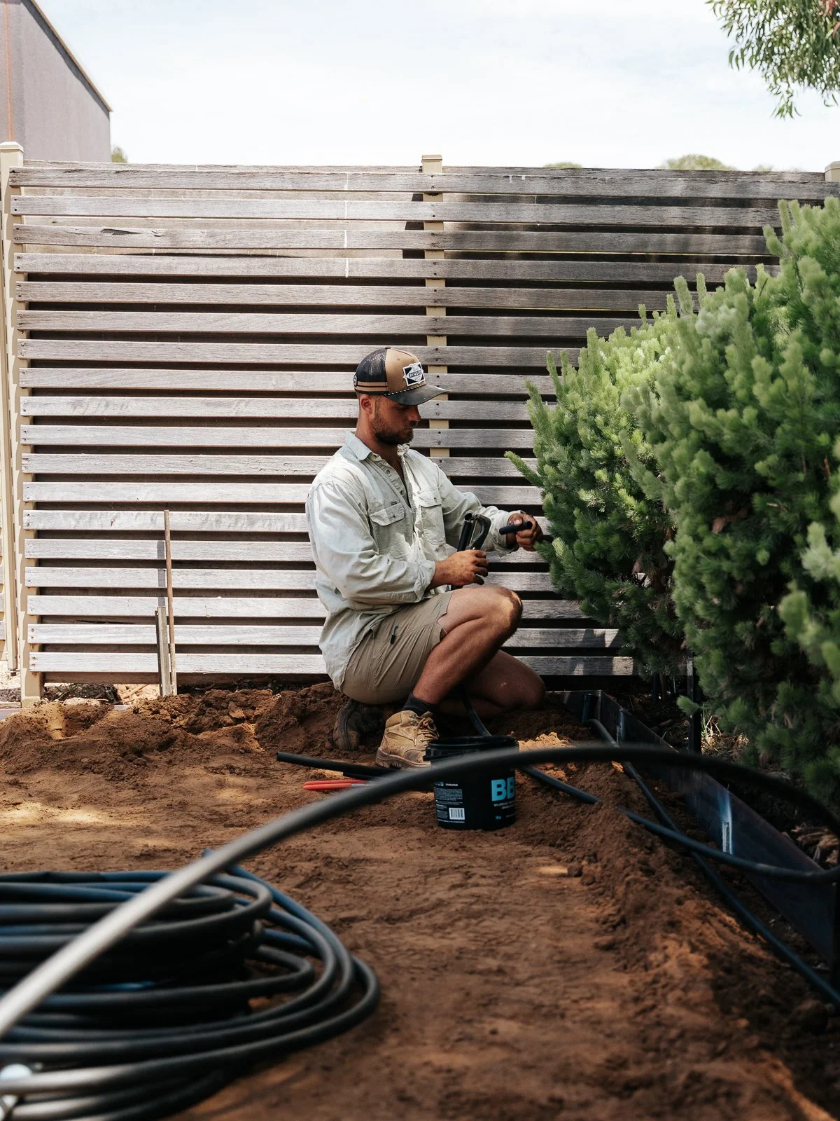 A man kneeling outdoors in a rural area, holding a small cylindrical device over a pile of soil, with soil and trees in the background.