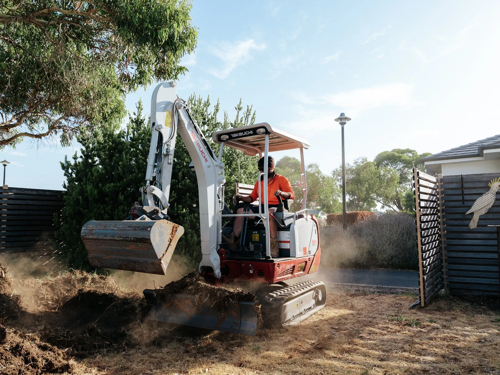 A person operating a small takeuchi excavator digging in a backyard next to a wooden fence and trees.