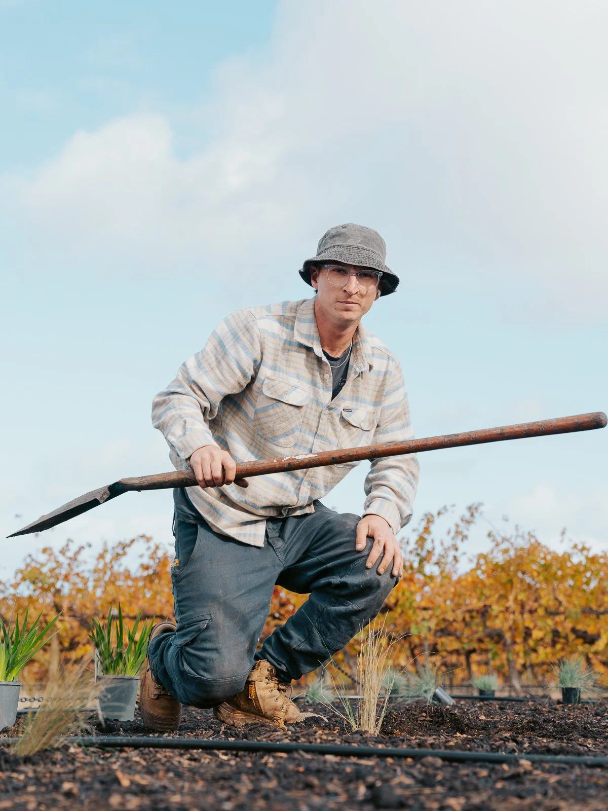 A man kneeling on the ground holding a gardening tool in a field with rows of plants and trees in the background.