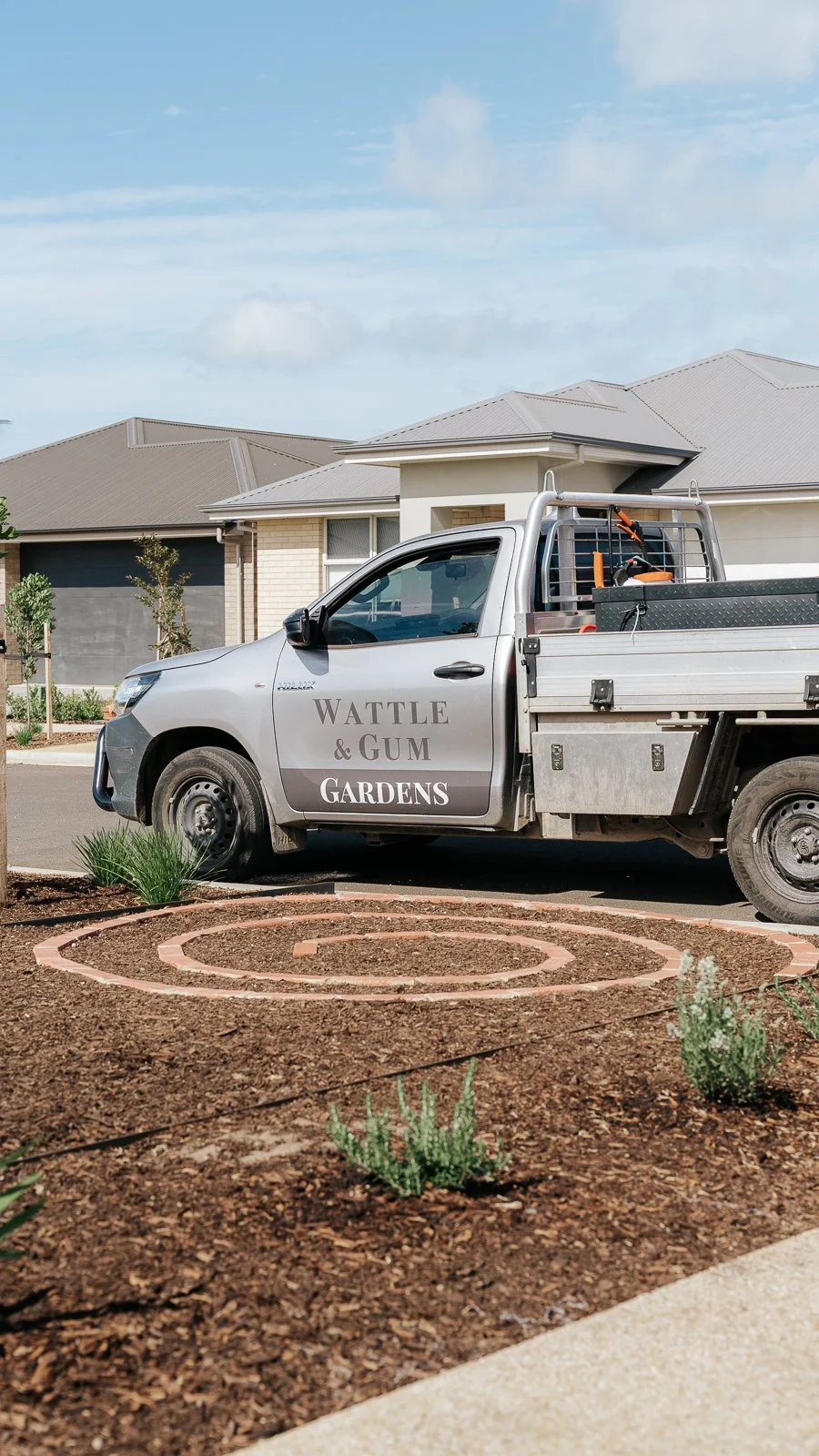 A pickup truck parked in a residential neighborhood with a sign on the door that reads 'Wattle & Gum Gardens.' The truck is parked next to a garden bed with circular brick edging and some small plants.