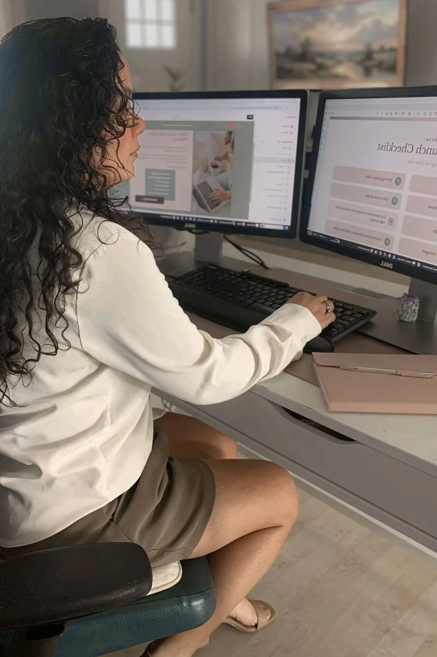 A woman with curly dark hair sitting at a desk with dual computer monitors, wearing a white blouse and shorts, with a pink notebook and pen on the desk, in a room with a painting on the wall.