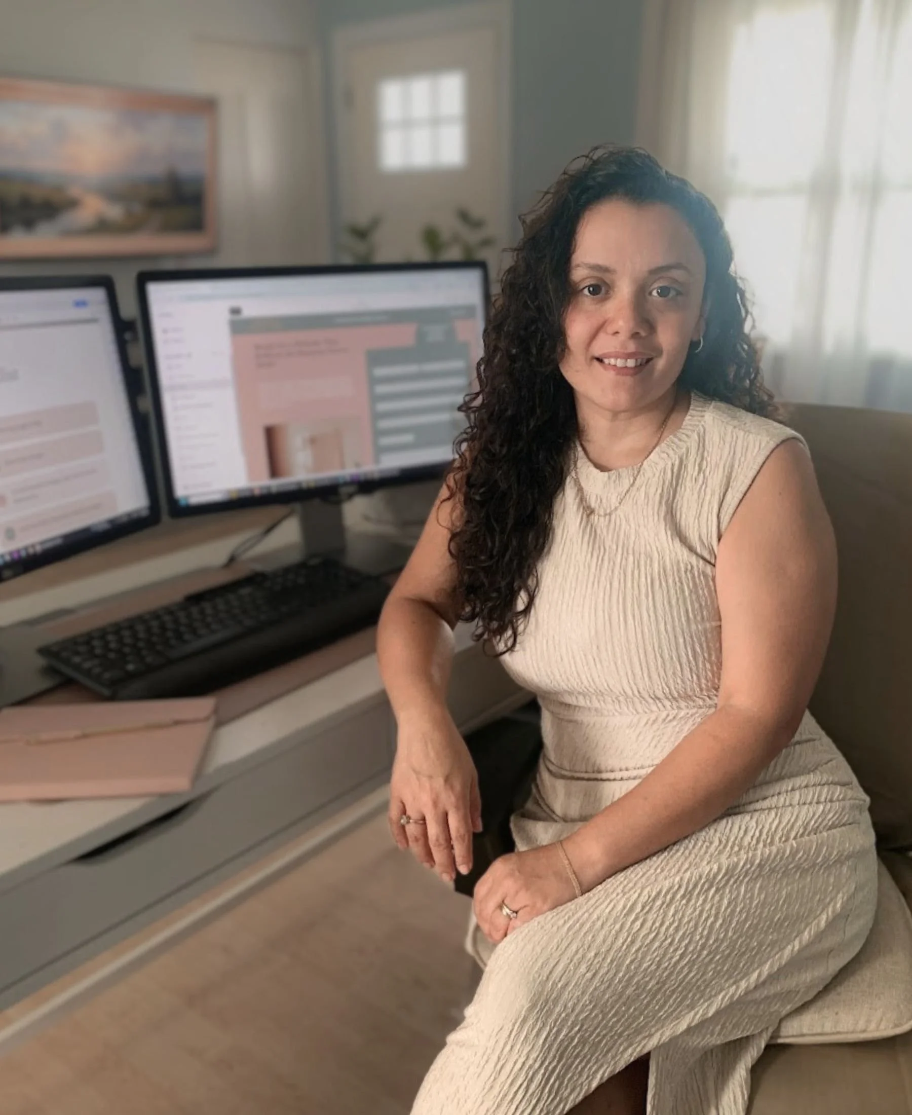 A woman with curly dark hair and a beige sleeveless dress sitting in front of a computer with three monitors in a room with natural light, a window with curtains, and framed artwork on the wall.