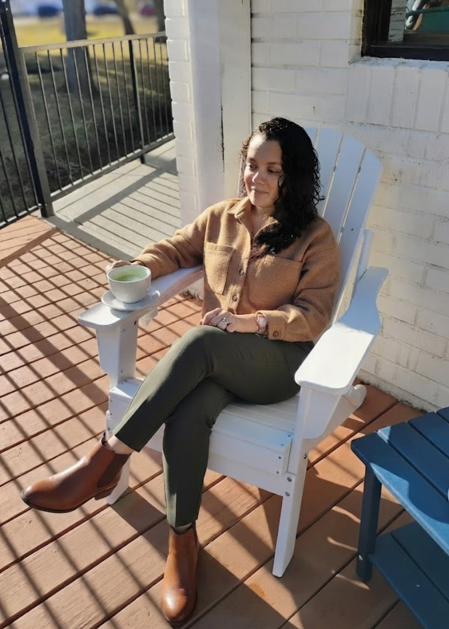 A woman sitting on a white outdoor chair, holding a cup of green tea, on a wooden deck outside a house with white brick walls and a window, wearing a tan coat, dark pants, and brown boots.