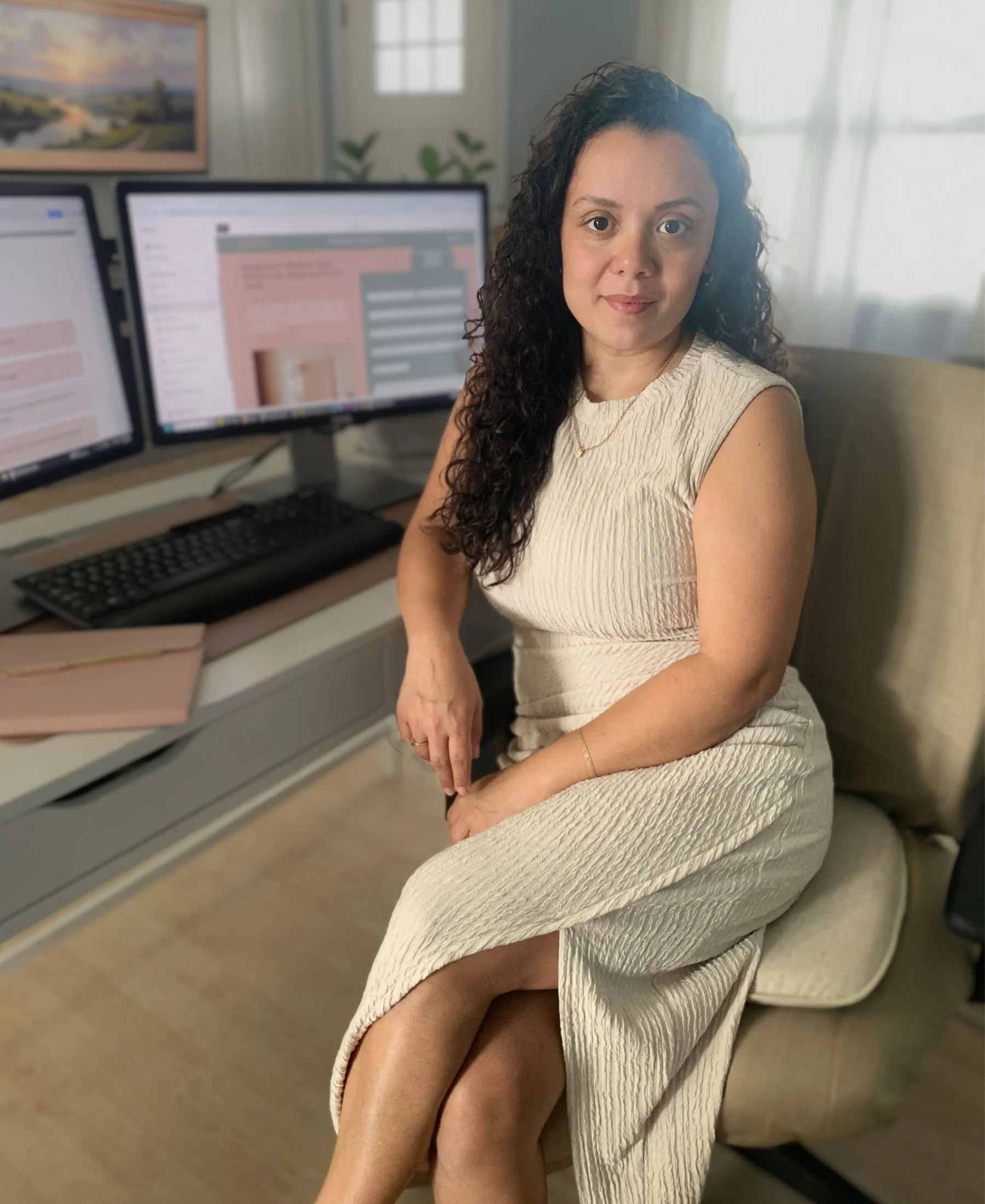 A woman with curly dark hair sitting on a beige armchair in a room with natural light, three computer monitors on a desk, a keyboard, and a keyboard tray, with a painting on the wall and window with curtains in the background.