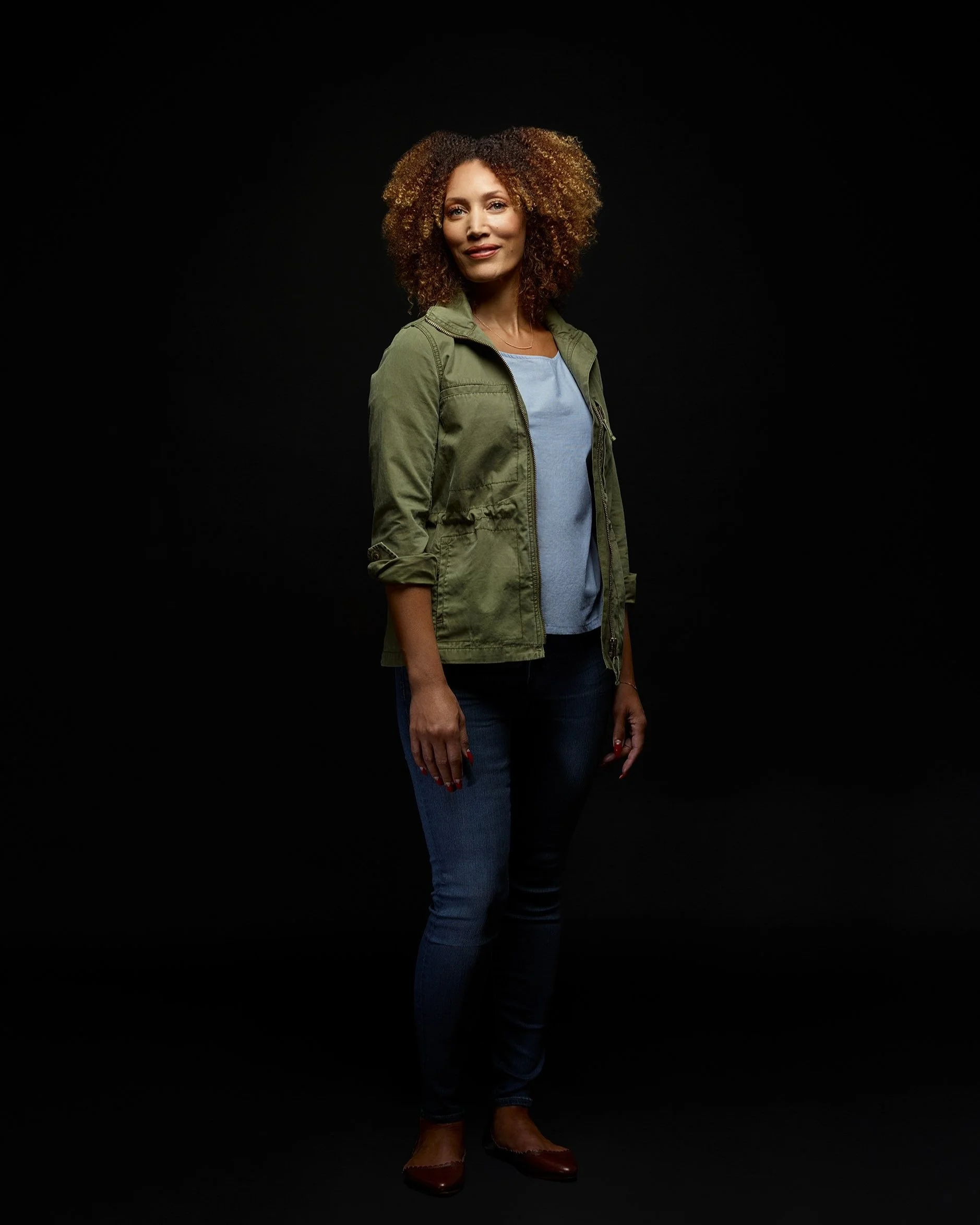Professional team headshot of woman in green jacket and jeans with relaxed studio pose