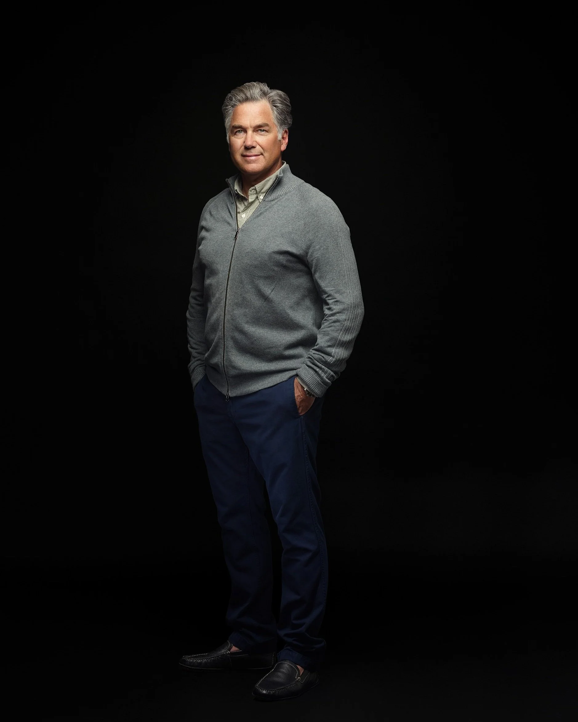 Team headshot of man in gray sweater and navy pants against dark studio backdrop