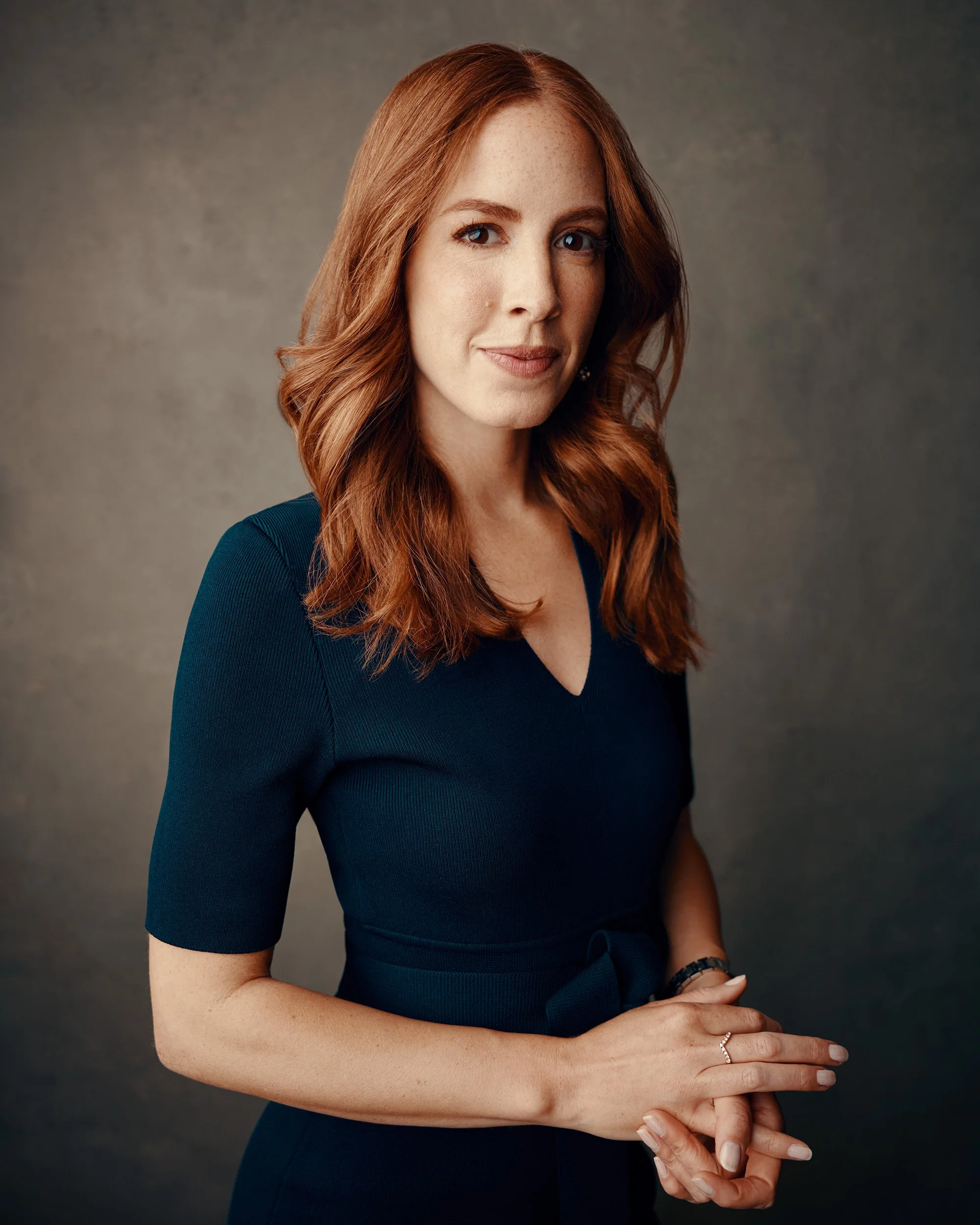 Company headshot of woman in navy dress with hands clasped against gray studio backdrop