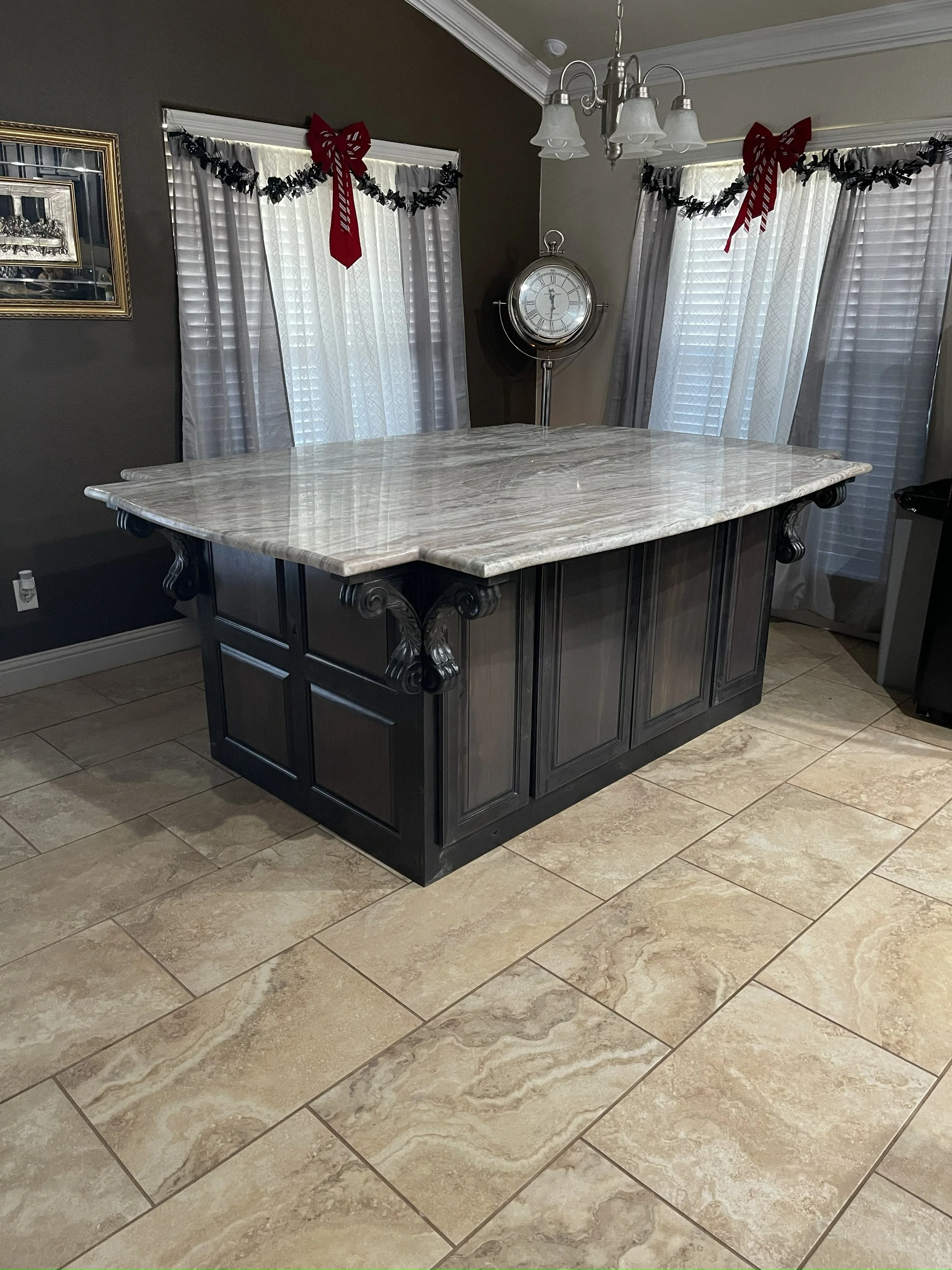 A kitchen island with a marble top and black cabinetry, situated on a tiled floor. Decorative garlands and red bows hang above the windows, and a silver clock is mounted on the wall behind the island.