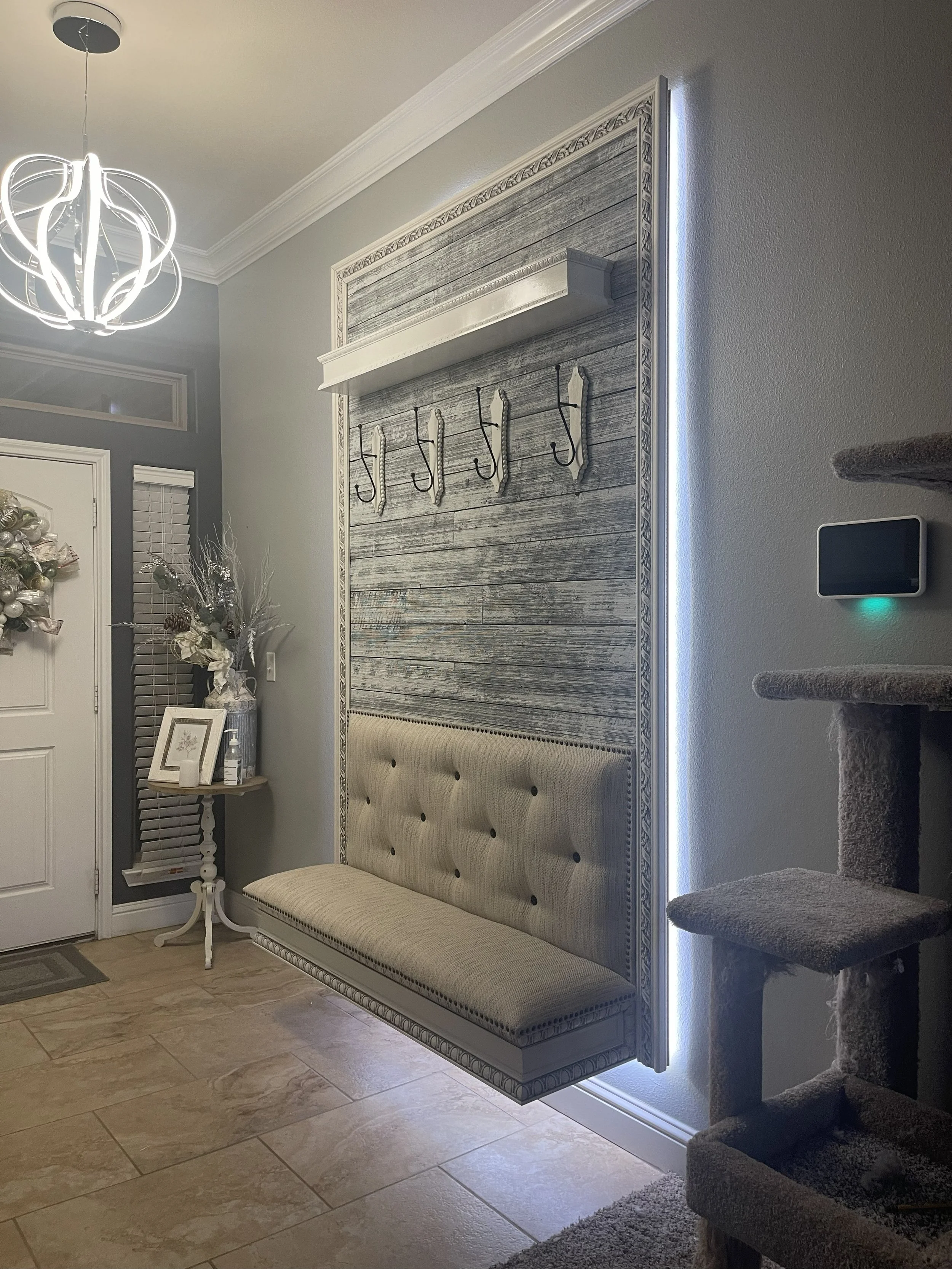 Interior of a home entryway with a decorative wall panel and coat hooks, a bench beneath the panel, a small table with framed pictures and a decorative arrangement, and a staircase with carpeted steps to the right. There is a front door with a Christ