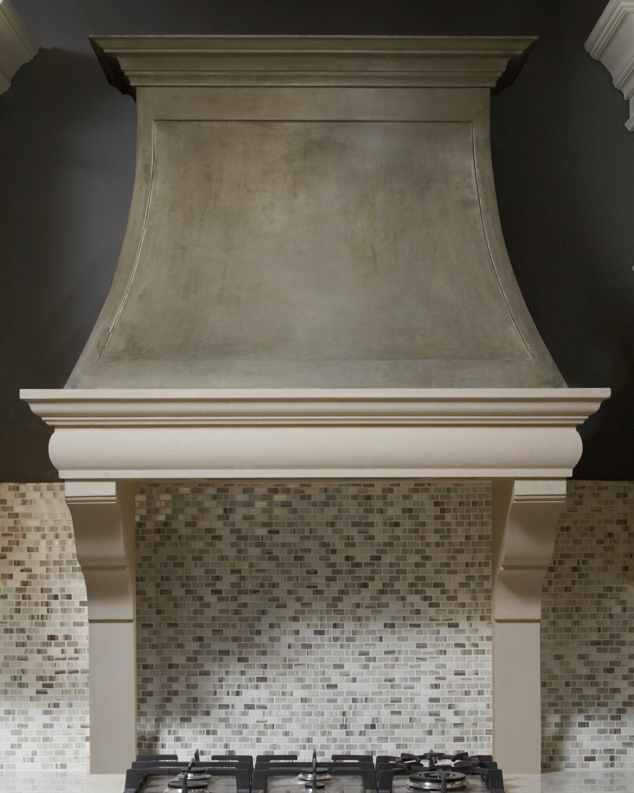 Kitchen range hood with beige ornate corbel supports above a gas cooktop, backsplash with small square mosaic tiles in neutral tones, wall painted dark charcoal, and crown molding.
