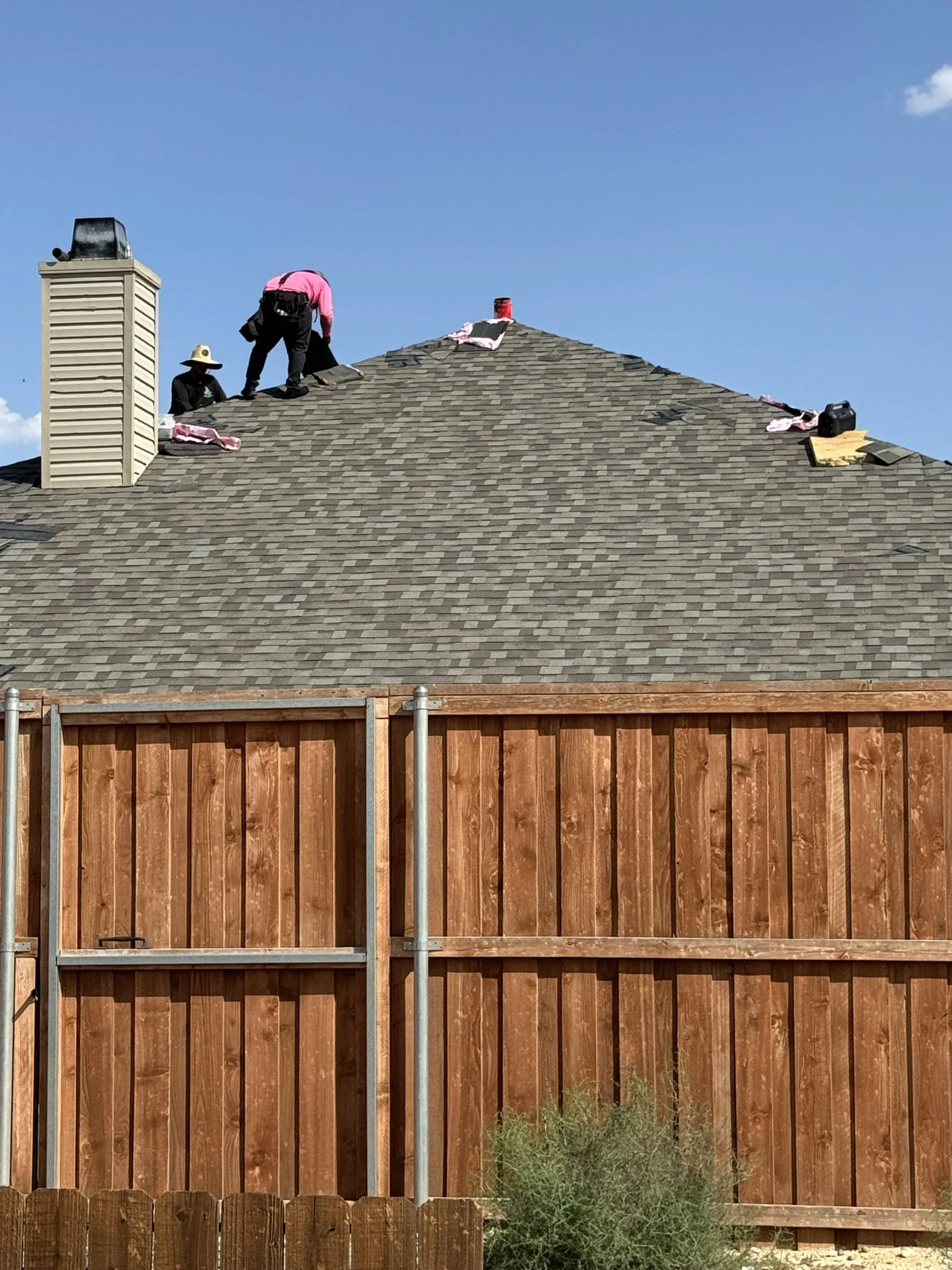 Two workers installing shingles on the roof of a house, with tools and materials nearby, under a clear blue sky.