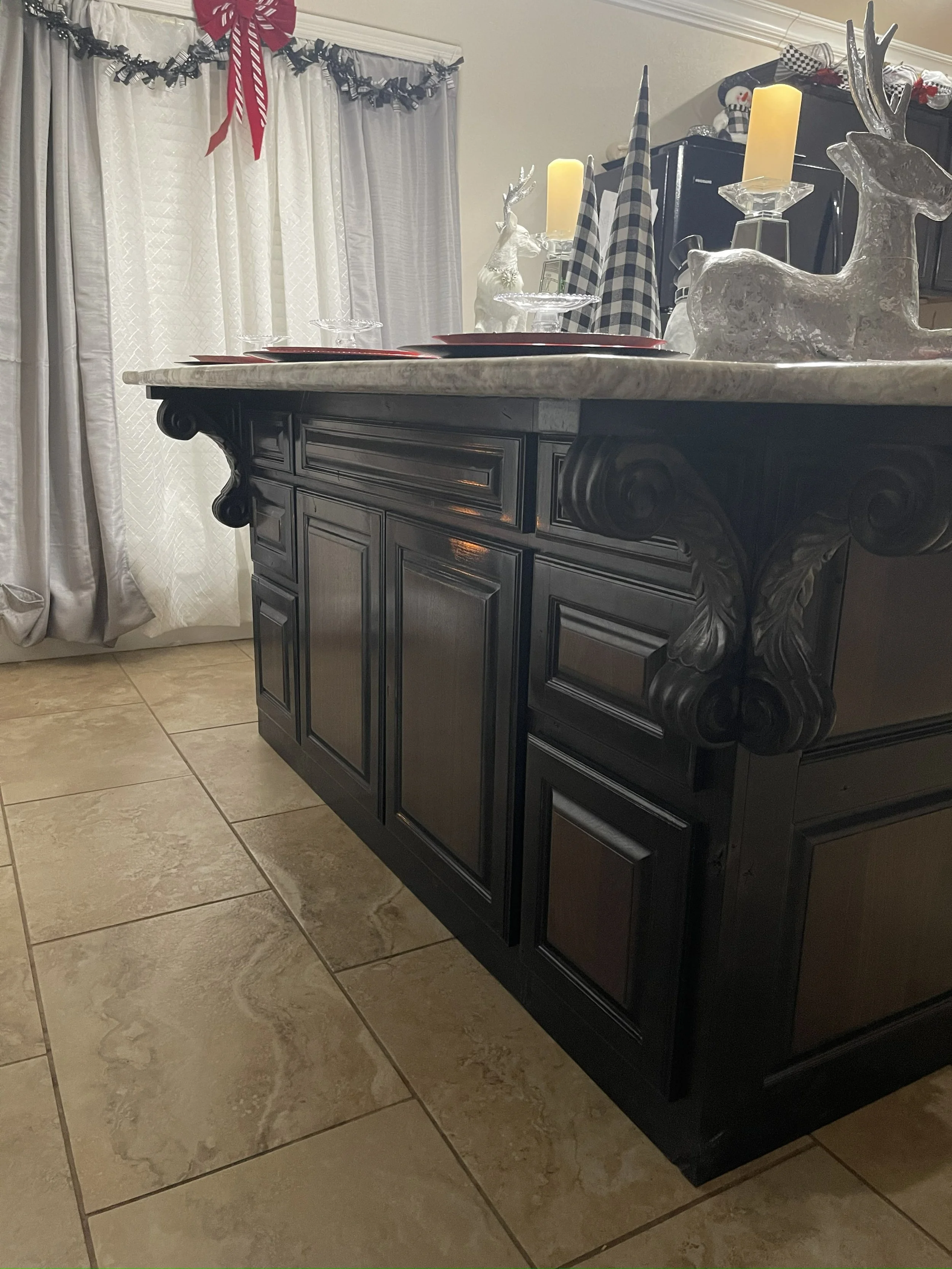 A black wooden sideboard with ornate carvings on its corner feet, topped with holiday decorations including reindeer figurines with candle holders, candles, and checkered fabric pieces. In the background, there are white curtains with a garland and b