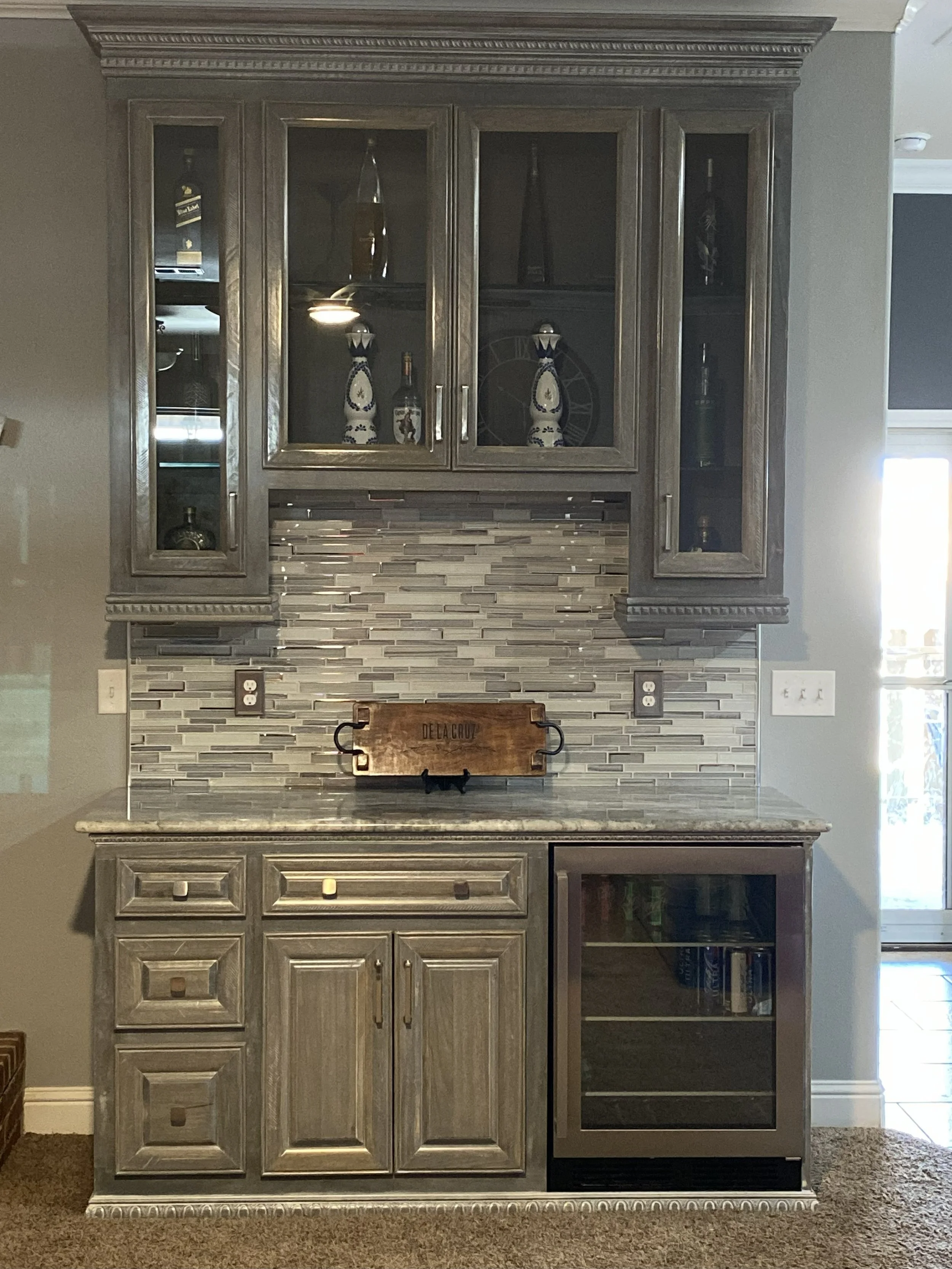 A gray wooden kitchen cabinet with glass doors on top, filled with bottles and decorative items, and closed doors below, set against a tiled backsplash and a gray wall.