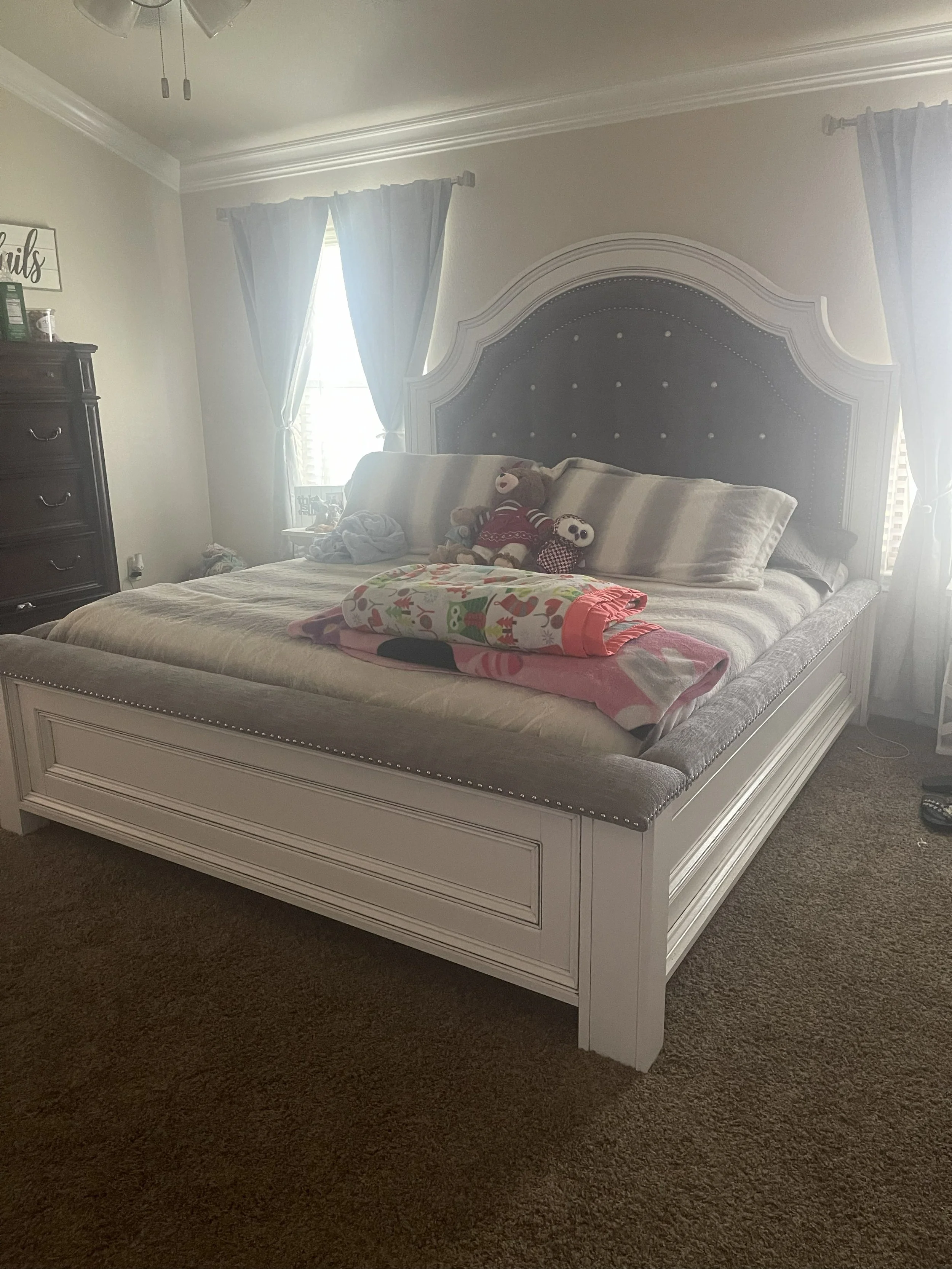 A bedroom with a white bed frame, beige bedding, and multiple pillows. There are three plush toys, including a teddy bear, on the bed. The room has beige walls, a window with blue curtains, and a dark wooden dresser on the left side. A holiday-themed