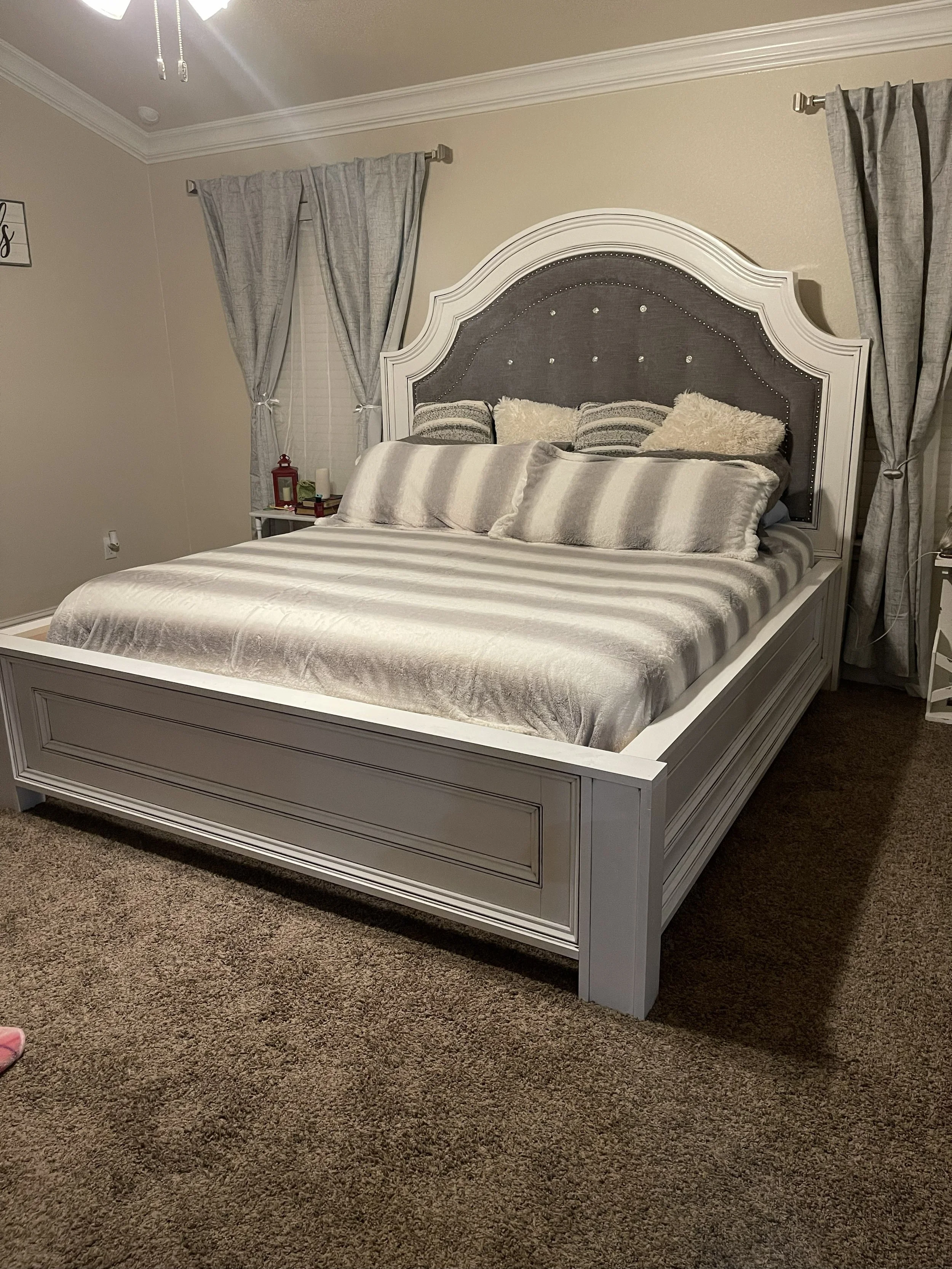 A bedroom with a large white wooden bed frame, gray tufted headboard with rhinestone accents, and plush pillows. There are gray curtains over the windows and a nearby nightstand with decorative items. The room has beige walls, crown molding, and a ce