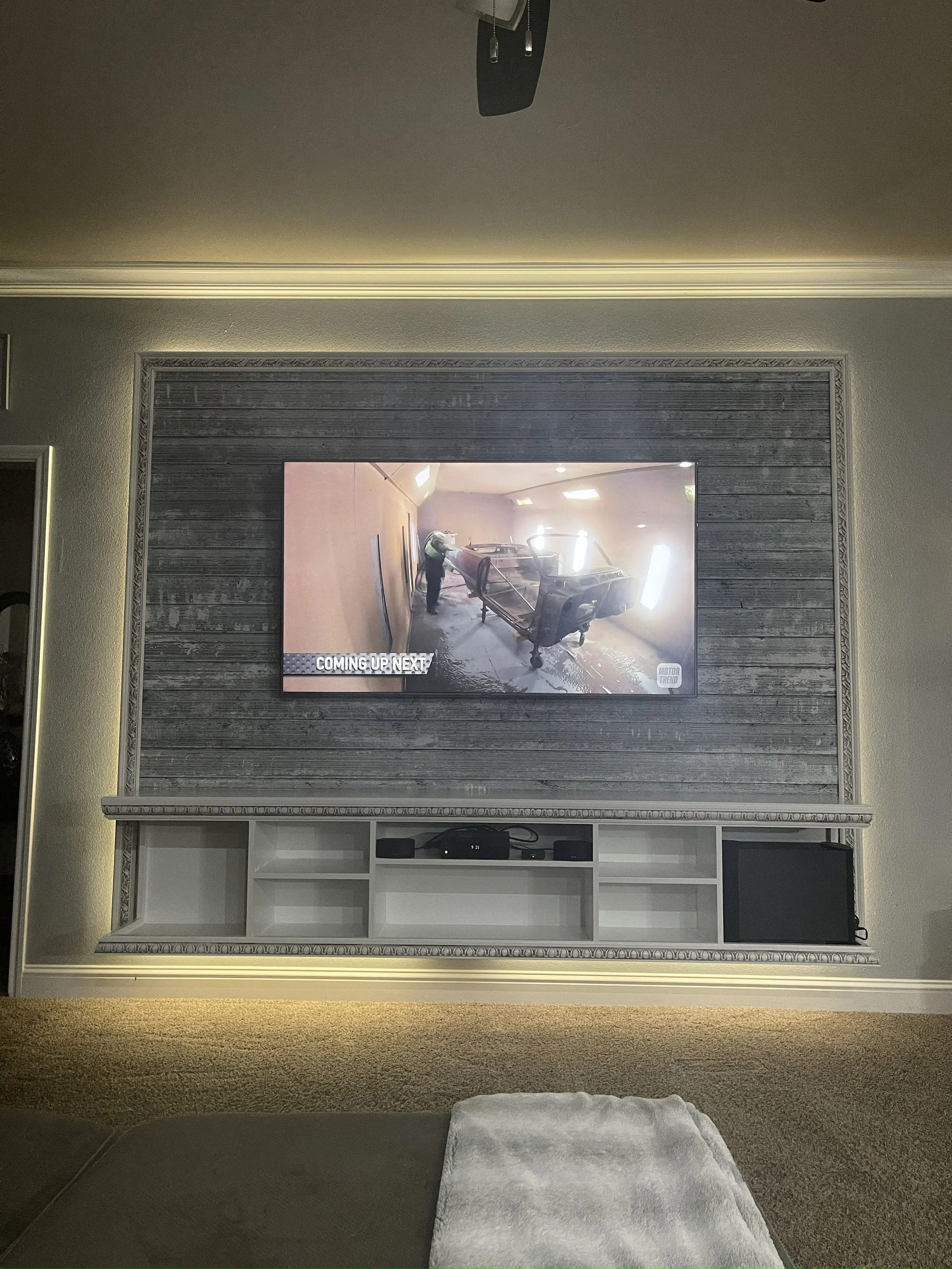 A living room with a large television mounted on the wall, displaying a scene with construction or workshop activity involving two workers and machinery. Below the TV, there is a white built-in shelving unit with multiple compartments, one containing