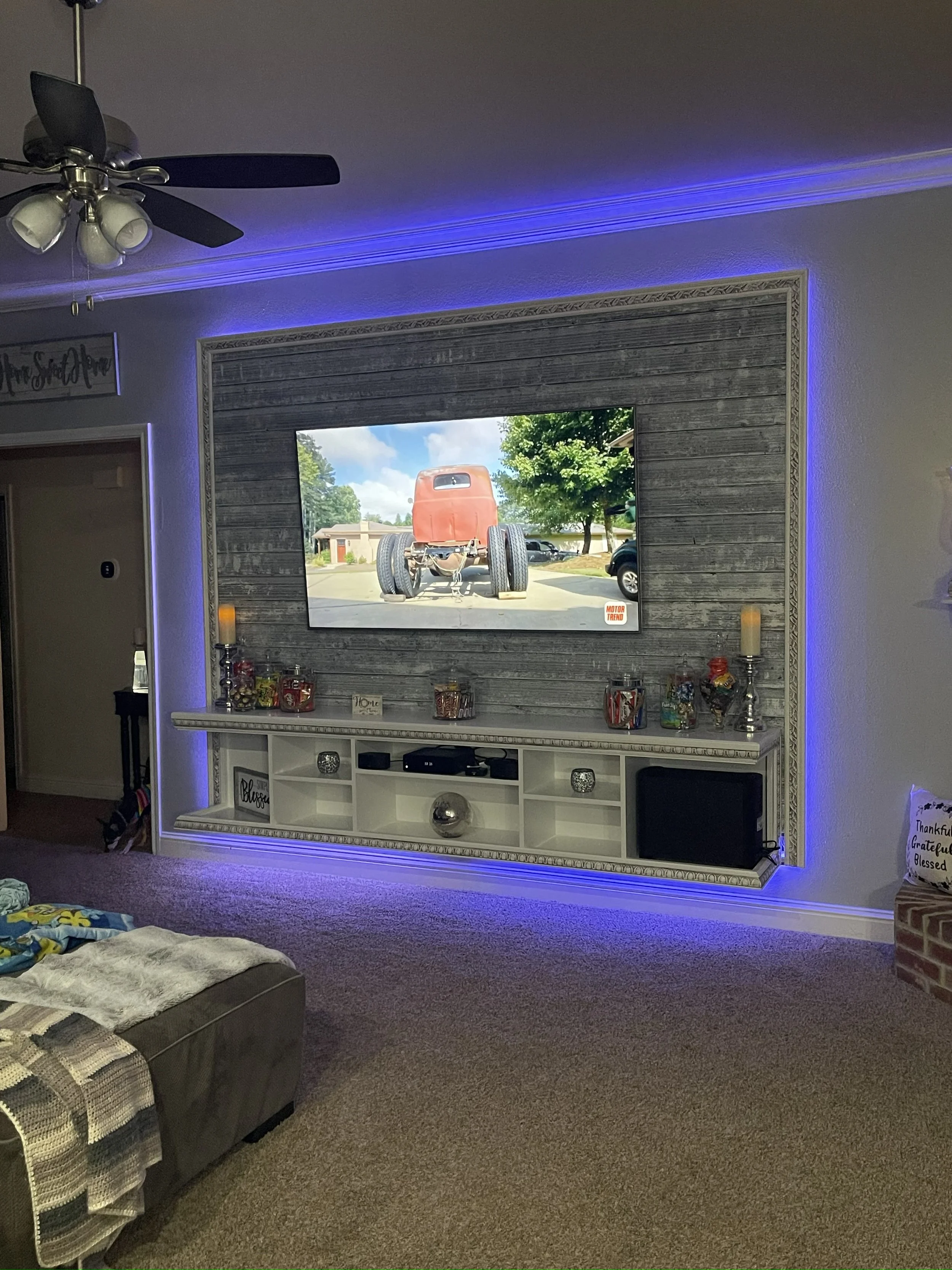 Living room with a large wall-mounted mirror outlined with LED strip lighting, a ceiling fan with lights, a TV, and a decorated shelf below the mirror. There's a partially visible couch with throw blankets in the foreground.