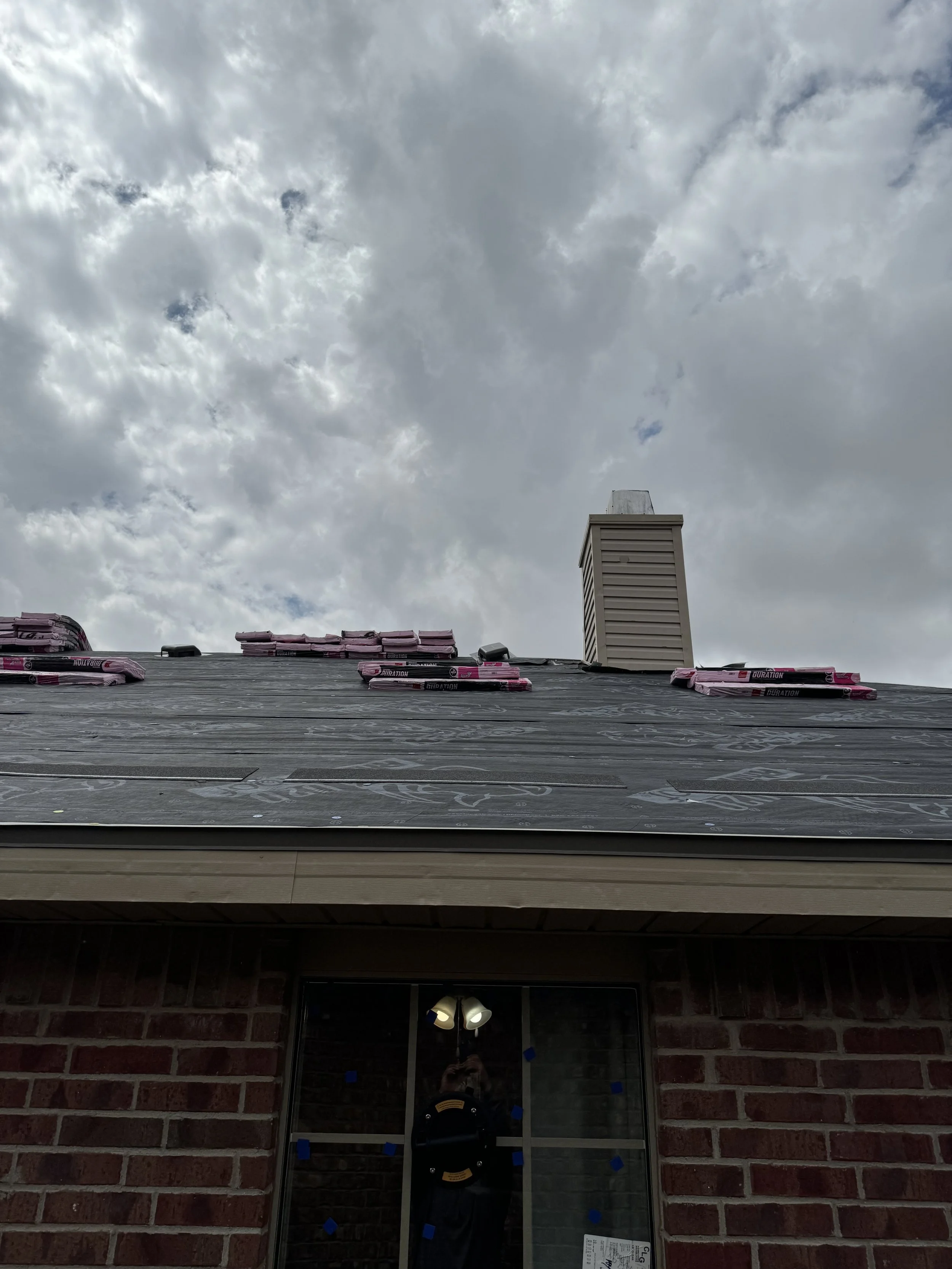 View of a house roof under construction with insulation layers, a brick wall, a window showing a reflection of a person, and a cloudy sky overhead.