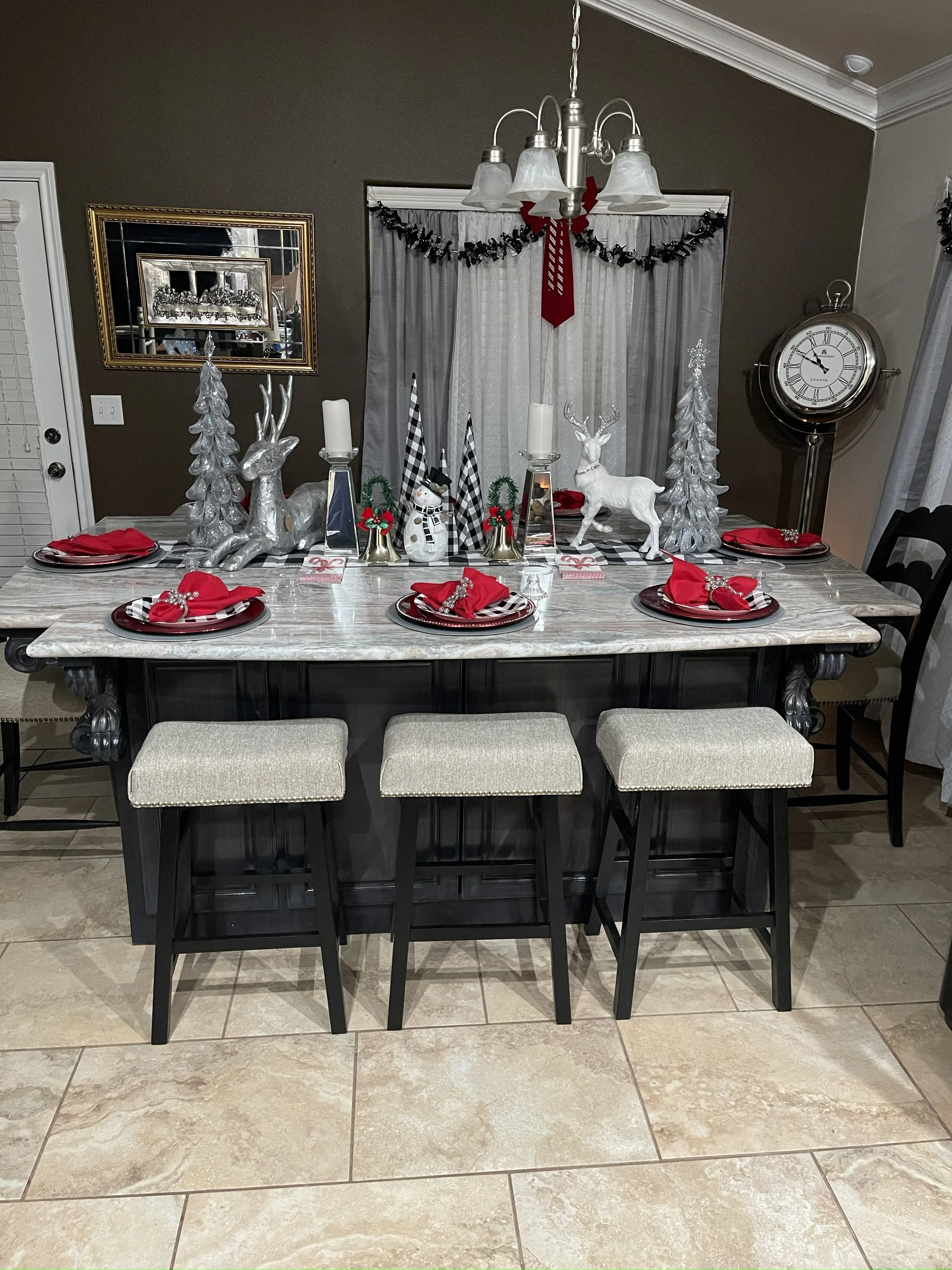 A dining table decorated for Christmas with red napkins, plaid plates, reindeer, Christmas trees, a snowman, candles, and a red and white tinsel garland. There is a large silver clock on the wall to the right, a mirror, and curtains with black garlan