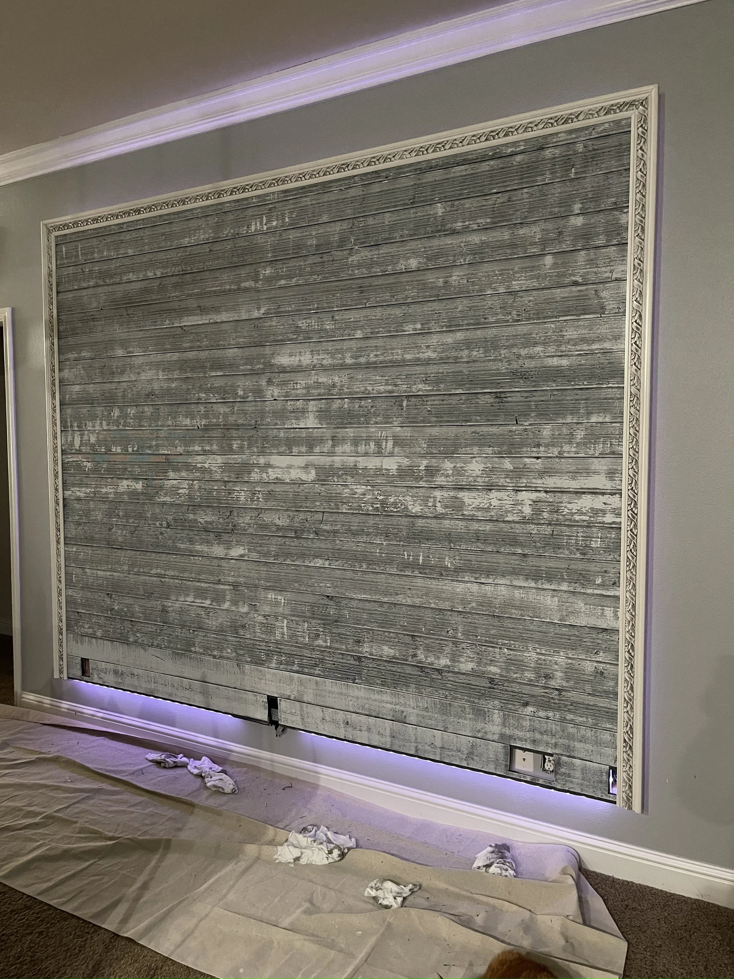 A decorated wall with a wooden textured panel, white ornate molding border, and purple LED lighting at the top, with a brown carpeted floor and painter's cloth and crumpled tissues on the ground.