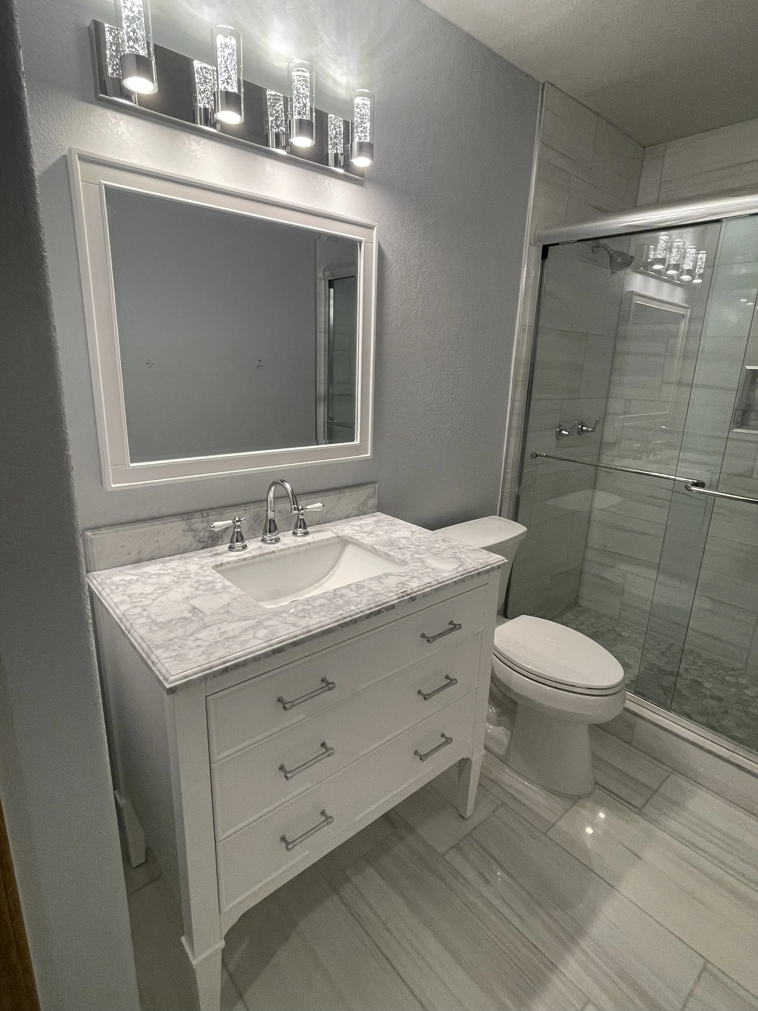A modern bathroom with a white vanity with a marble countertop, a mirror above, a sleek faucet, a toilet, and a glass-enclosed shower with tiled walls.