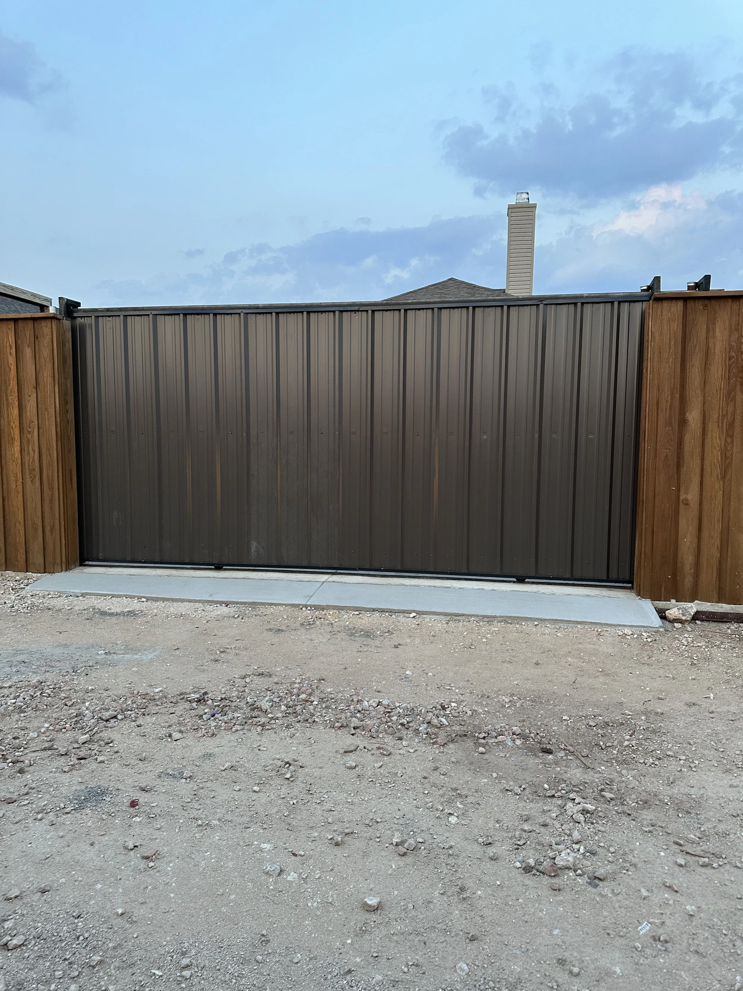 A backyard with a metal gate and wooden fencing, dirt ground, and a cloudy sky with scattered clouds.