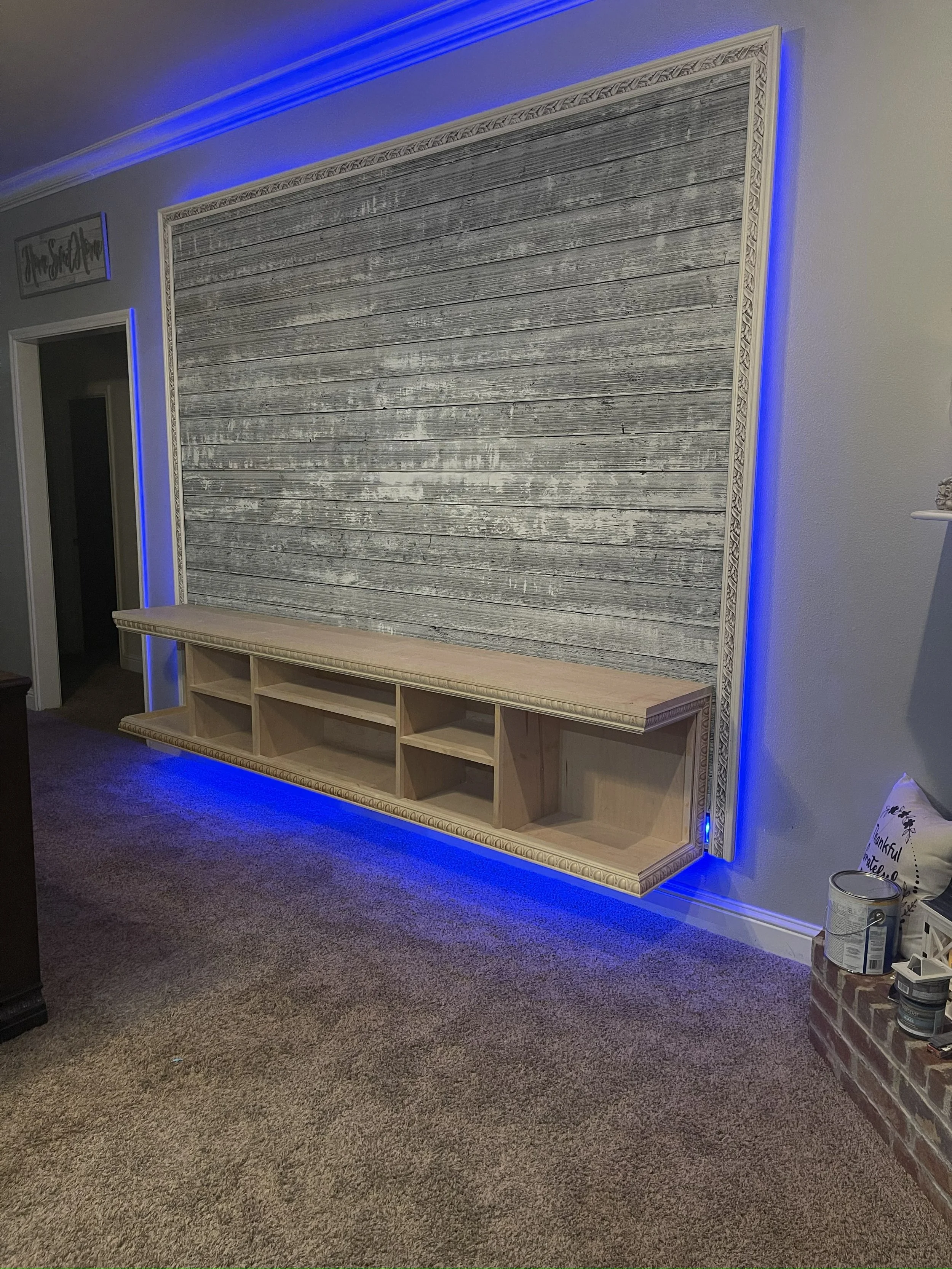 Living room with a large wall decoration made of horizontal weathered wood planks, framed with ornate wood border and accented with blue LED lighting along the top and bottom edges. There is a wooden entertainment console below the decoration and a c