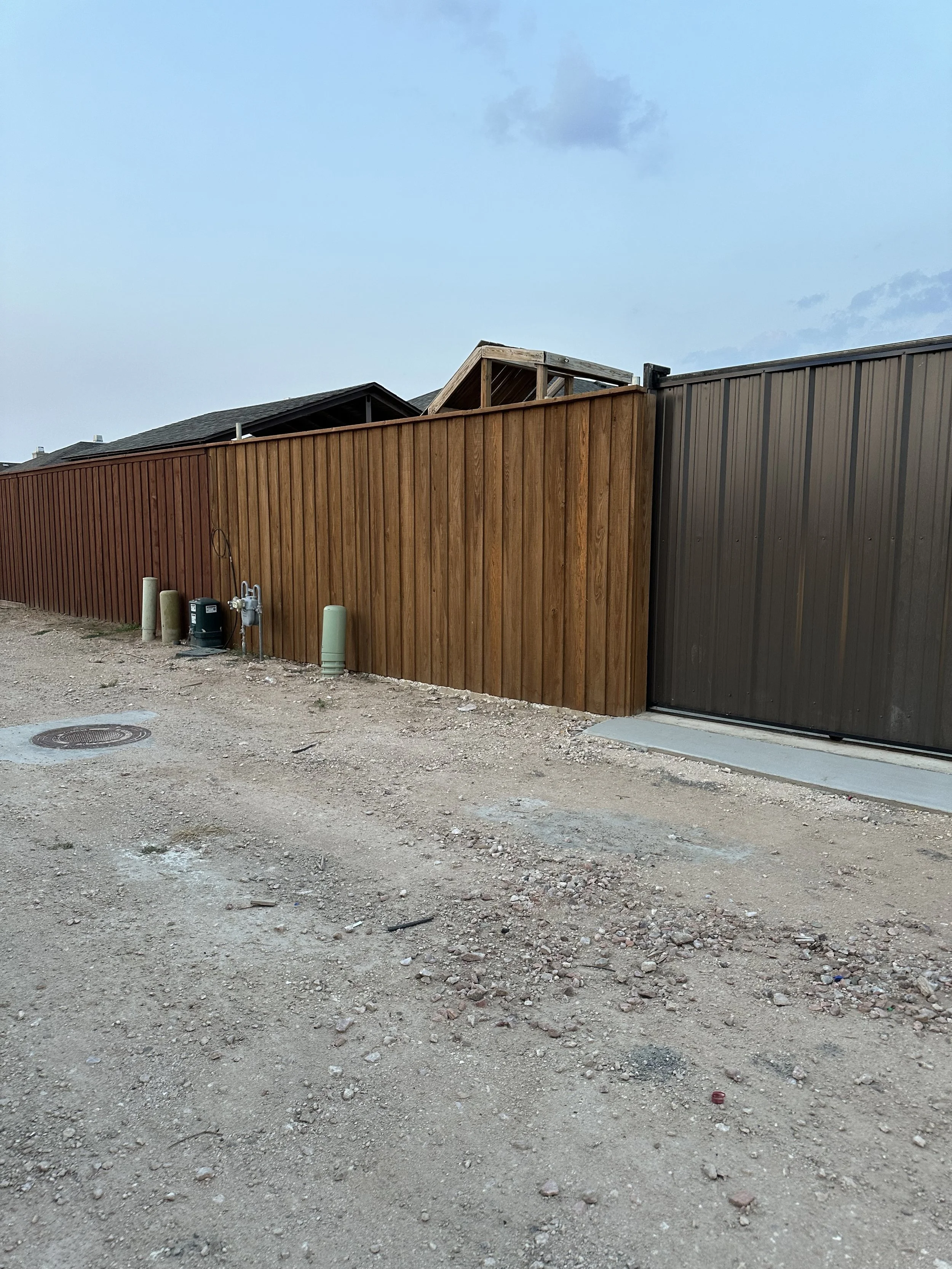 A dirt lot with scattered rocks and debris in front of a tall wooden fence, with a house roof visible behind the fence and some utility pipes and fittings near the fence.