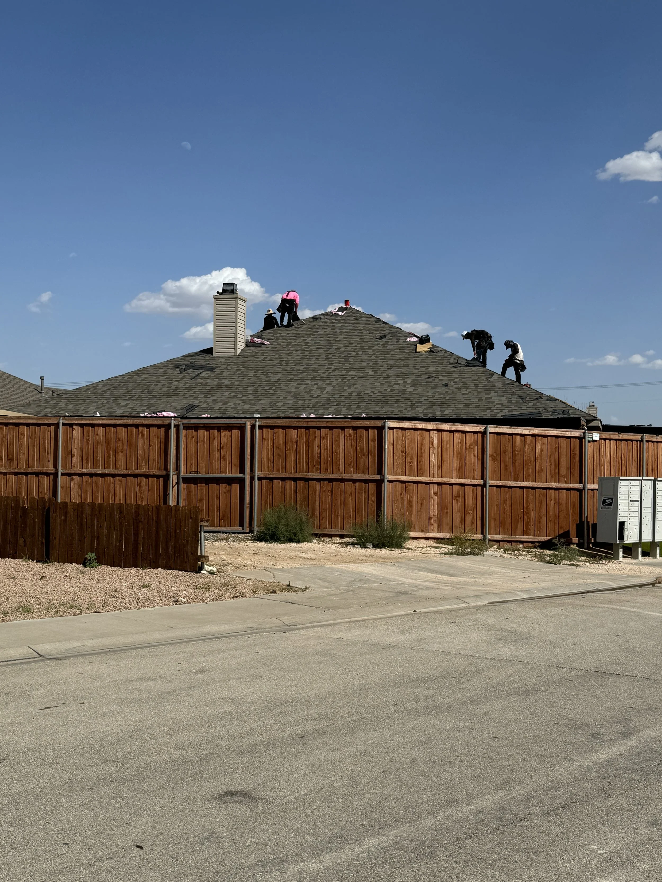 People working on roof of house during sunny day, fenced backyard, sidewalk and street in foreground, blue sky with a few clouds and visible moon in background.