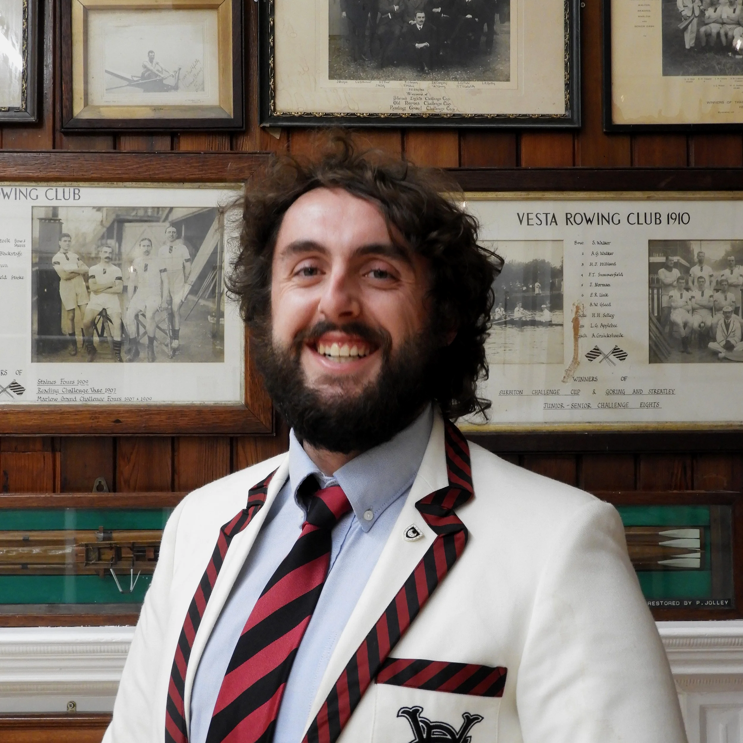 Smiling man with dark curly hair and beard, wearing a white blazer, red and black striped tie, and white shirt, standing in front of framed vintage rowing team photographs on wood-paneled wall.