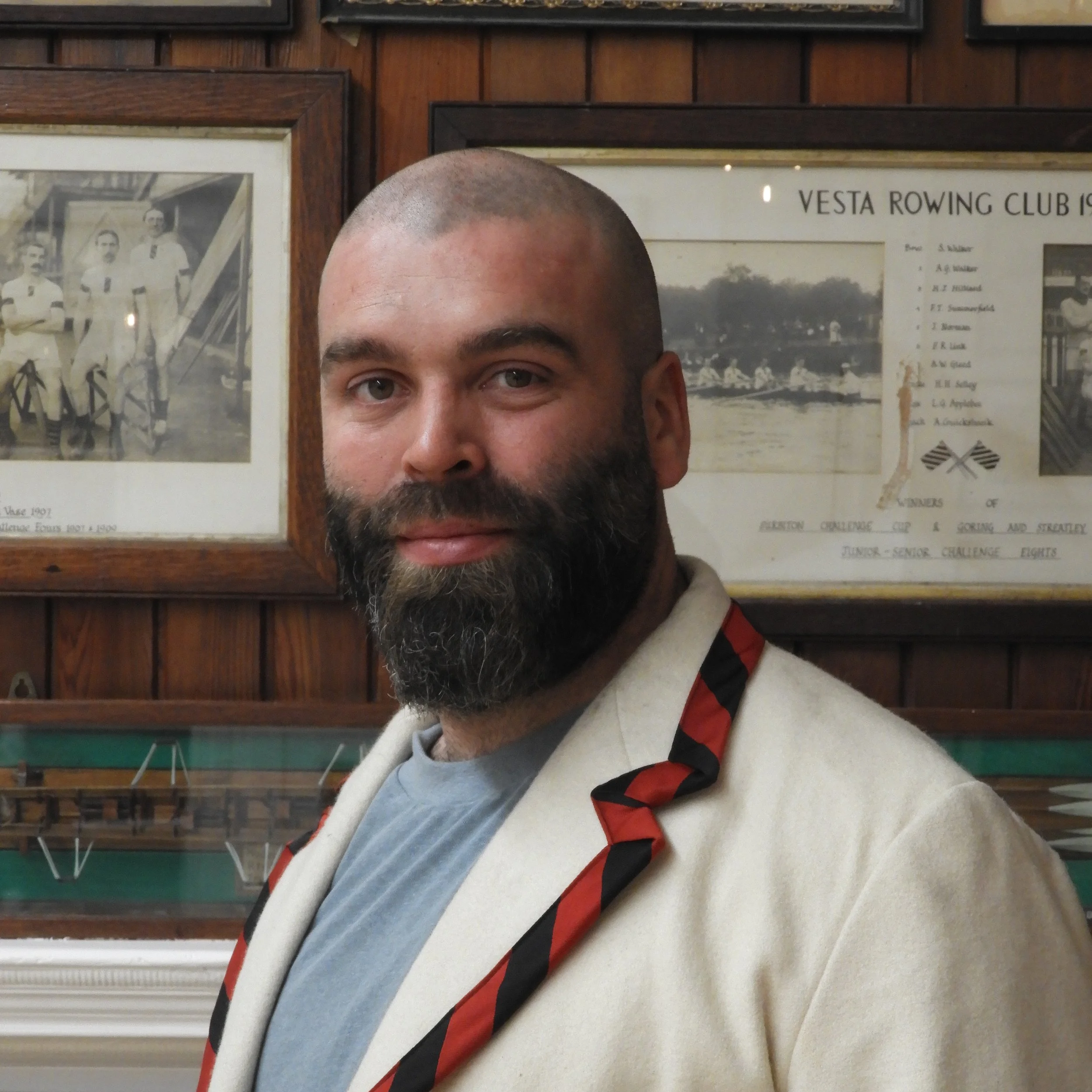 Close-up of a man with a shaved head and beard, wearing a white shirt and a black, red, and white striped tie, standing in front of framed photographs and posters on a wooden wall.