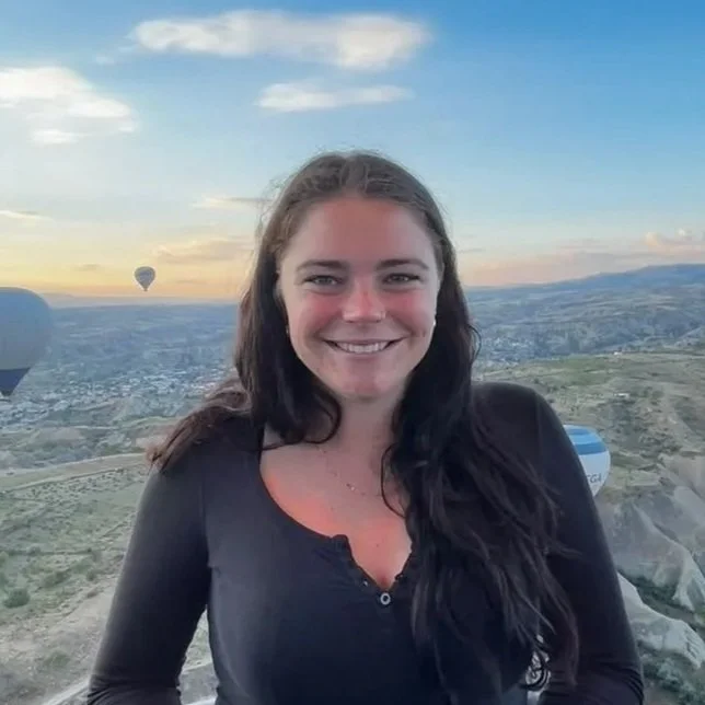 Young woman smiling at sunset with hot air balloons in the sky and a landscape below.
