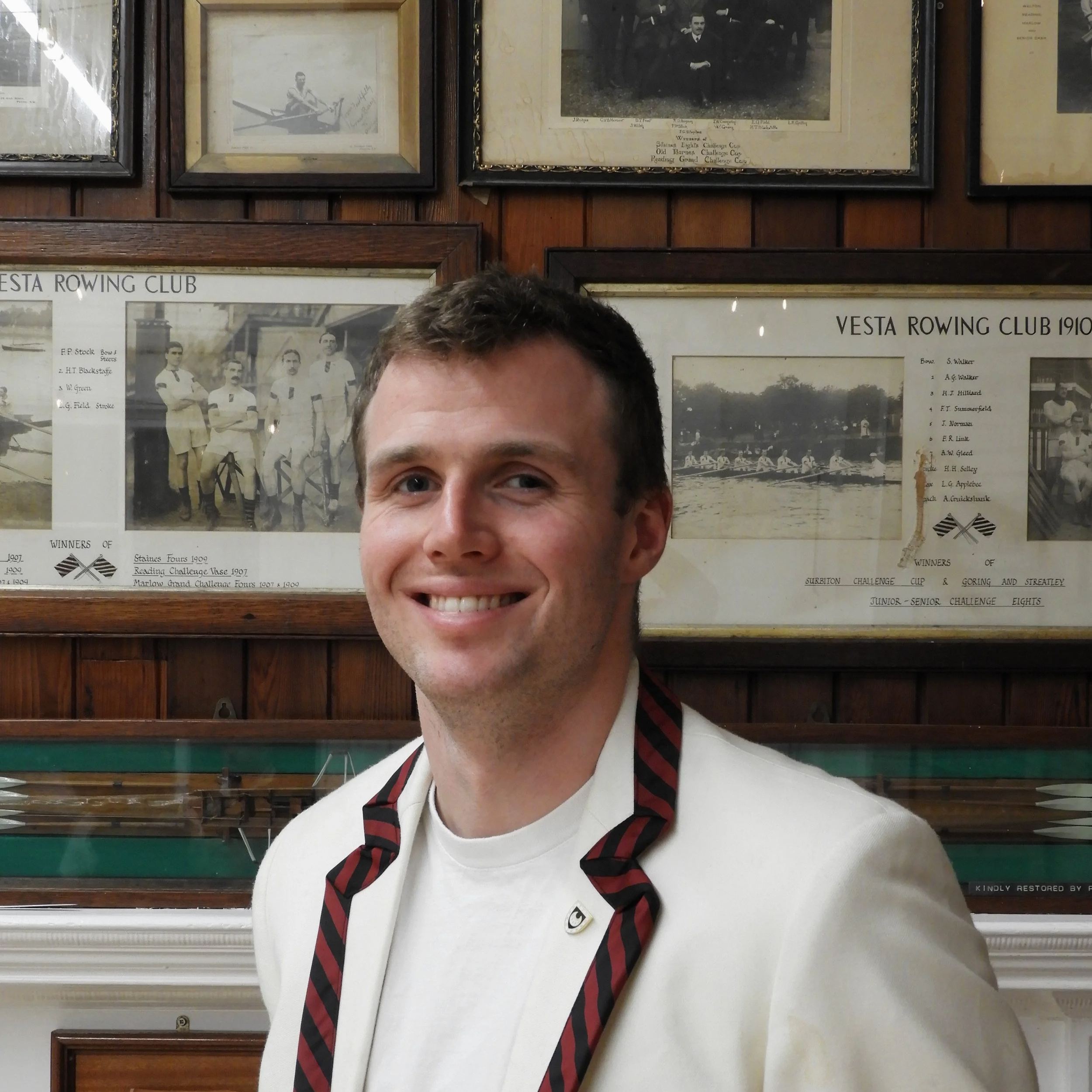 A young man wearing a white shirt, black and red striped tie, and a white blazer takes a selfie indoors. The background features framed historic black-and-white photographs and posters on a wooden wall, related to rowing clubs and competitions.