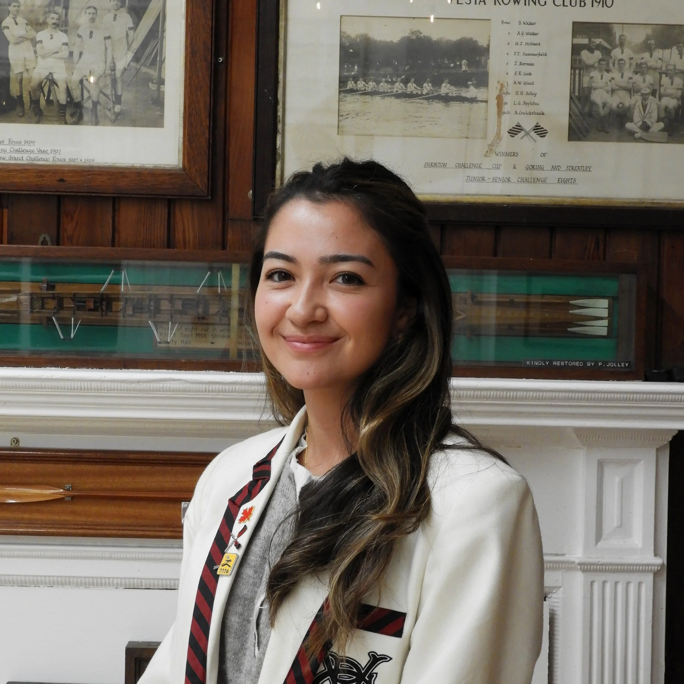 A woman with dark hair and a white coat standing in front of framed photographs and documents on a wooden wall, including a history of a rowing club.