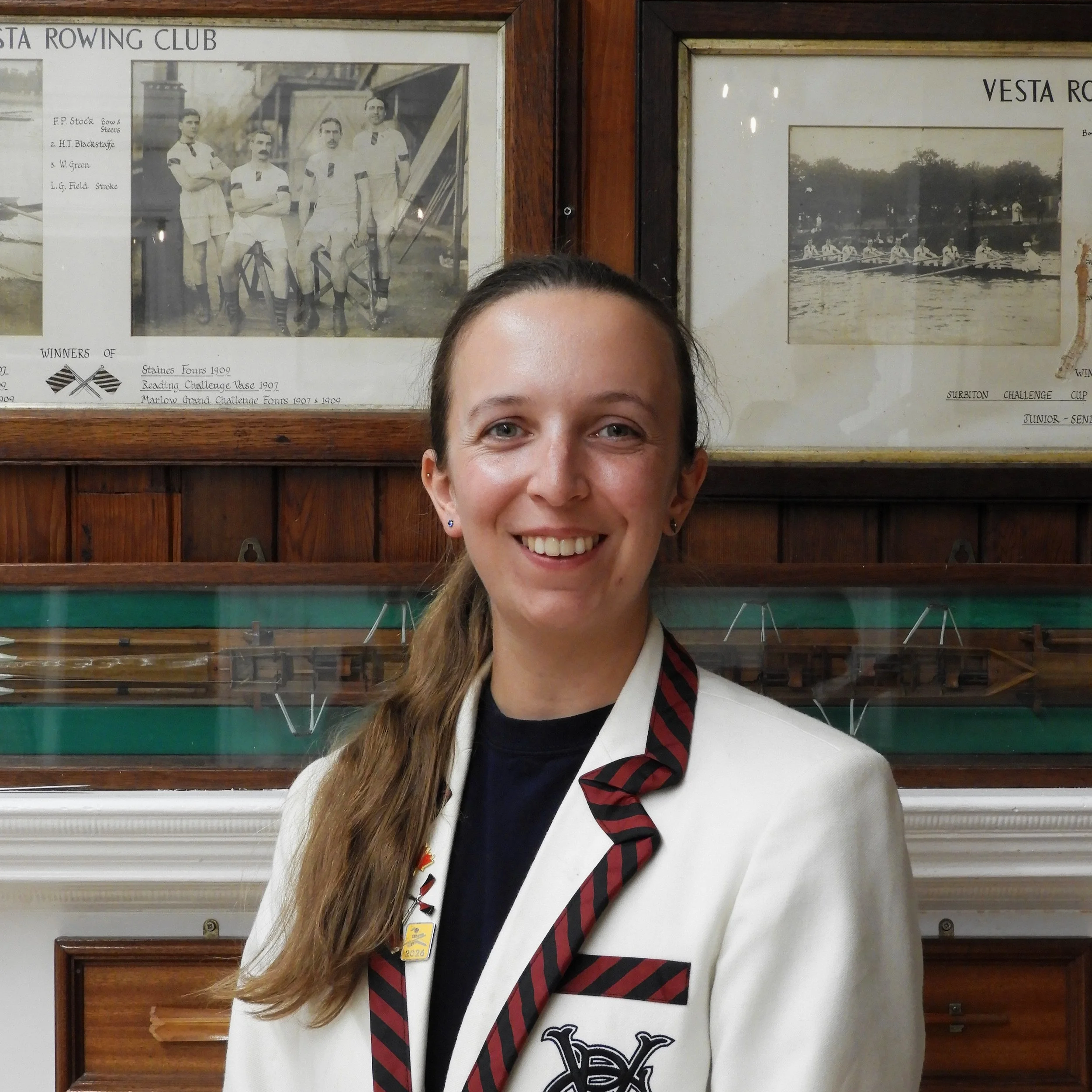 A smiling young woman with long brown hair, wearing a white blazer and a black and red striped tie, standing in front of framed black-and-white historic photographs of rowing teams on a wooden wall.