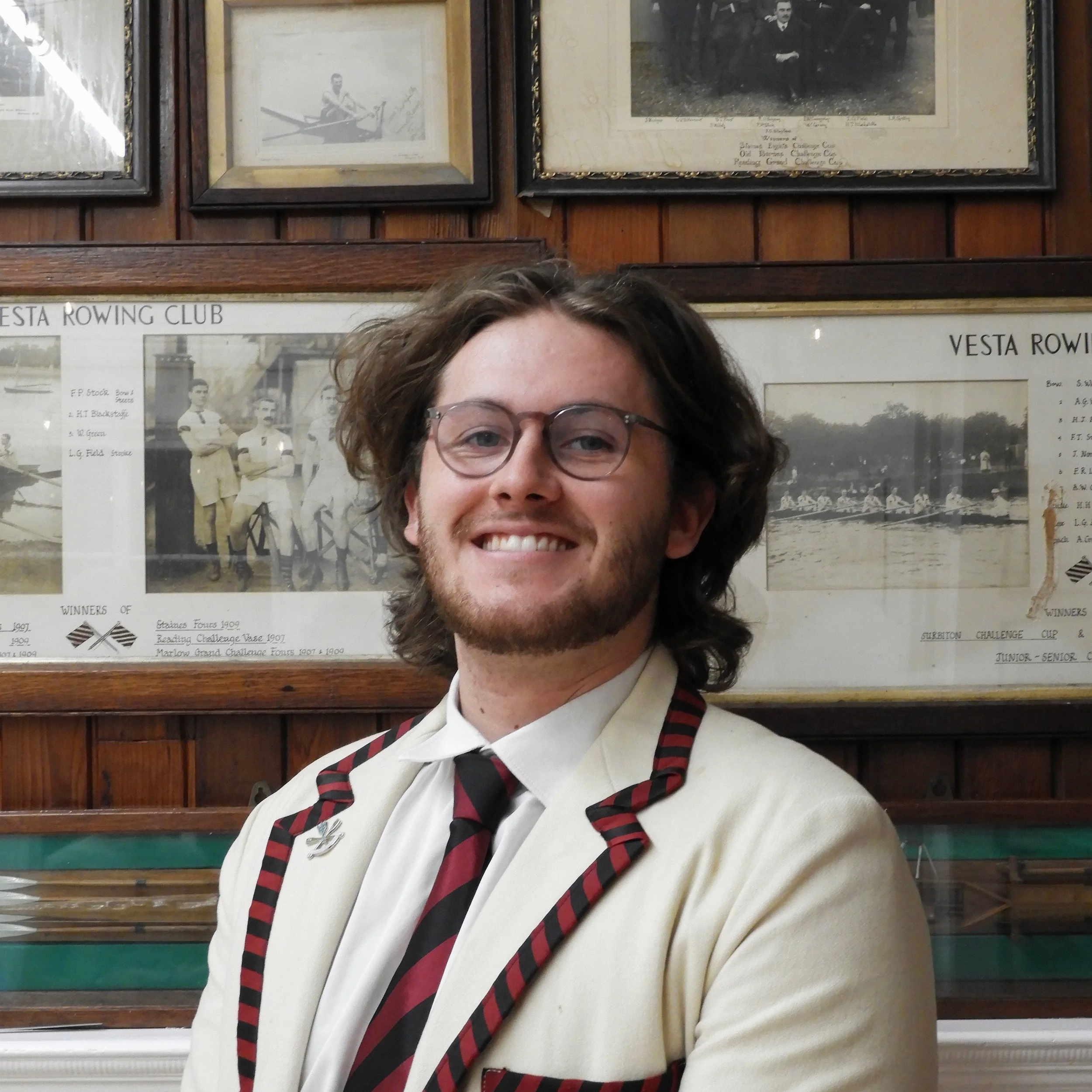 A person with curly brown hair, glasses, and a white sweater with red and black plaid accents, smiling and standing in front of framed vintage black and white photographs on a wooden wall.