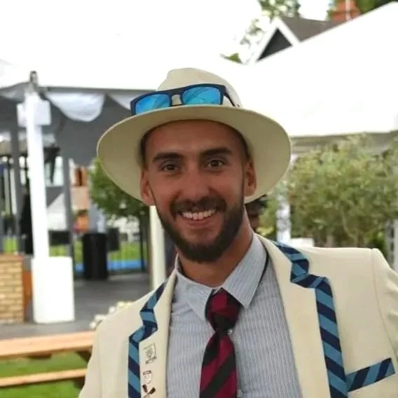 Man smiling outdoors, wearing a white hat with blue sunglasses, a light blazer with blue accents, a striped shirt, and a dark tie.