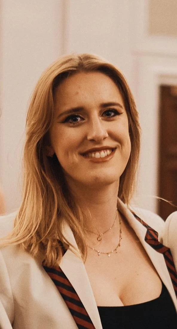 A young woman with shoulder-length blonde hair smiling, wearing a white blazer with striped accents and layered necklaces, indoors with a blurred background.