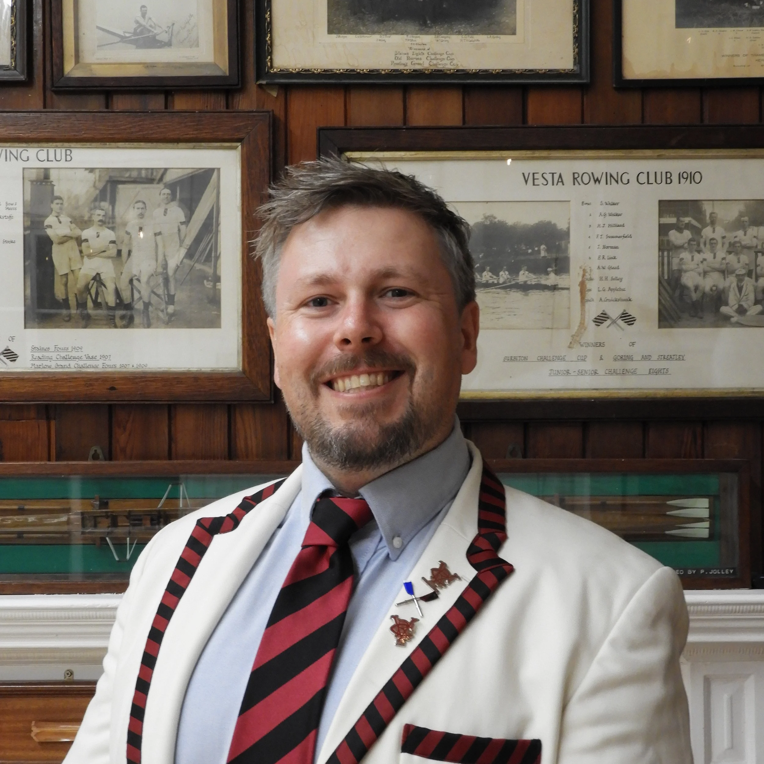 A man with short, gray hair and a big smile taking a selfie in front of a wall of framed historical photographs and documents about rowing clubs.