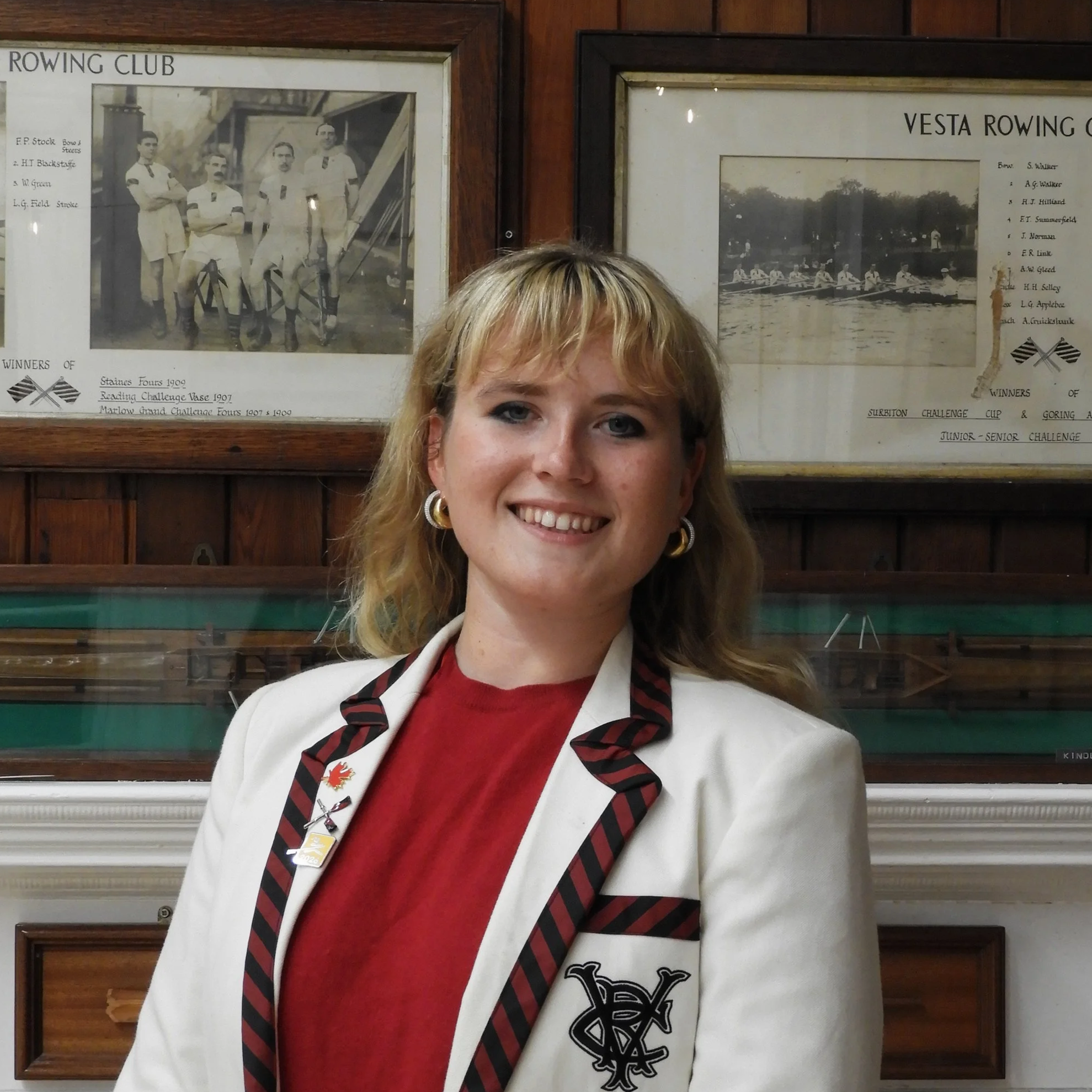 A woman with blonde hair and earrings smiling in front of framed vintage rowing team and club photos on a wooden wall.