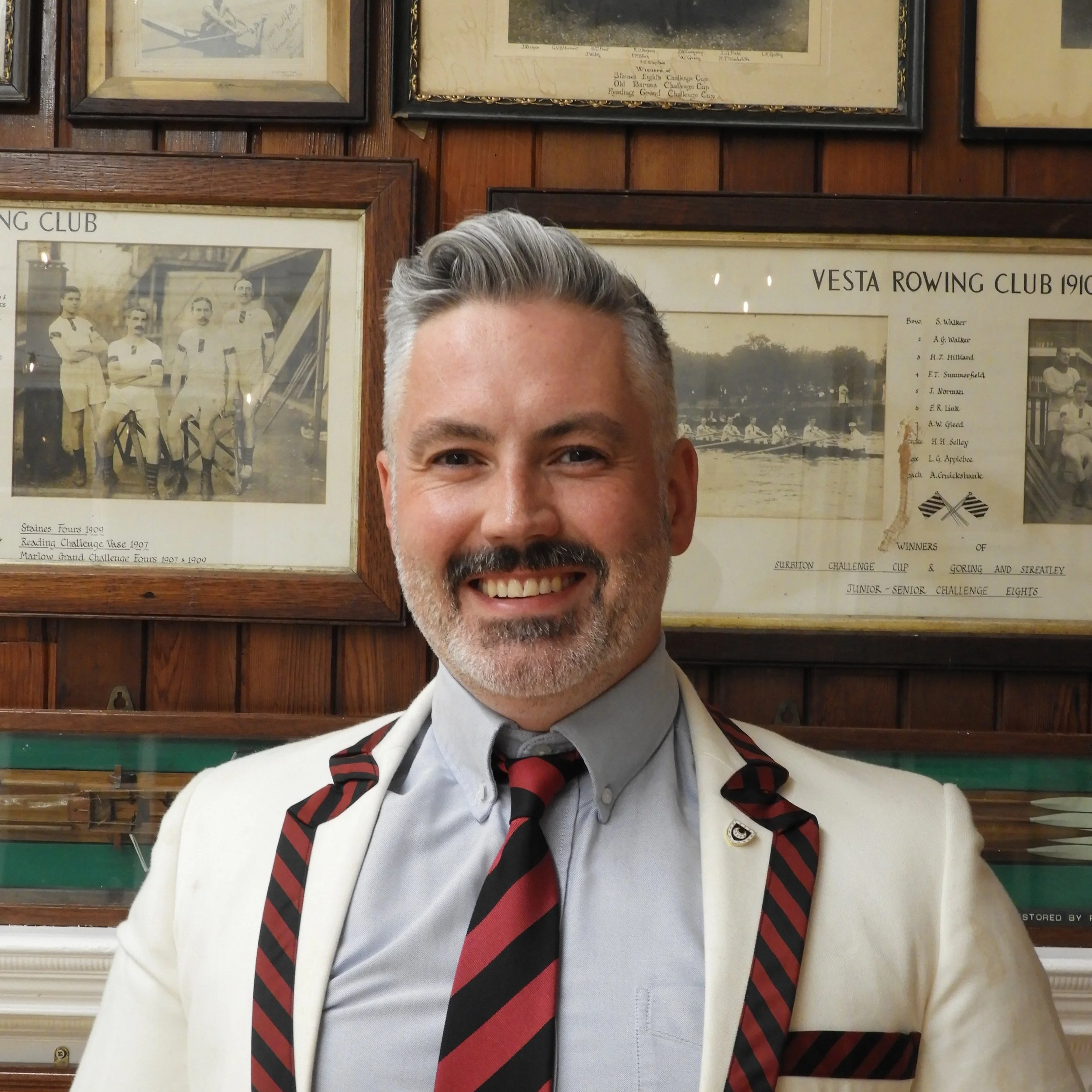 Head and upper torso of a middle-aged man with gray hair and beard, smiling, wearing a white blazer, gray shirt, and a black, red, and gray striped tie, standing in front of a wooden wall with framed vintage sports and rowing club photographs and plaques.