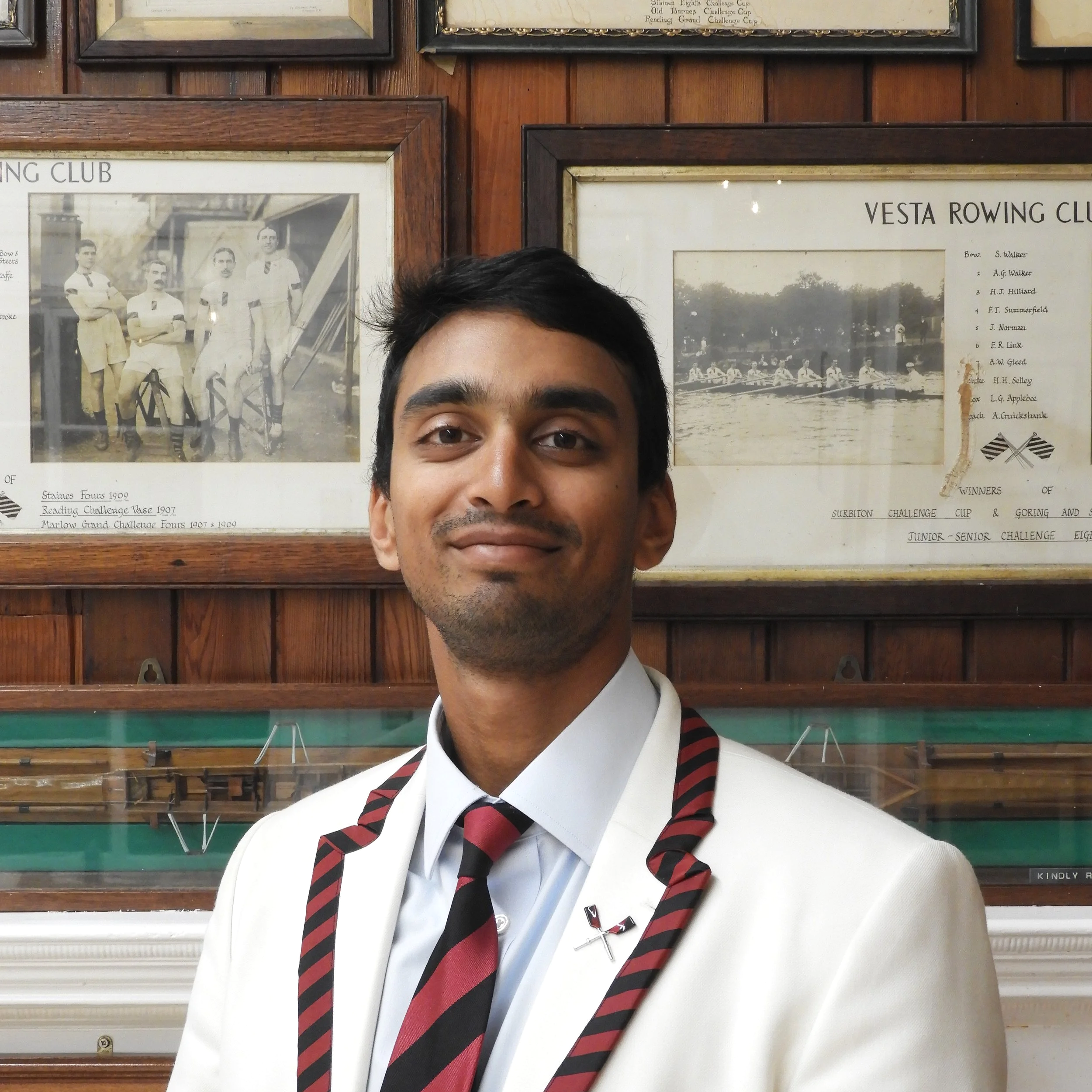 A person's face and upper body partially visible in front of framed rowing team photographs and awards on a wooden wall.