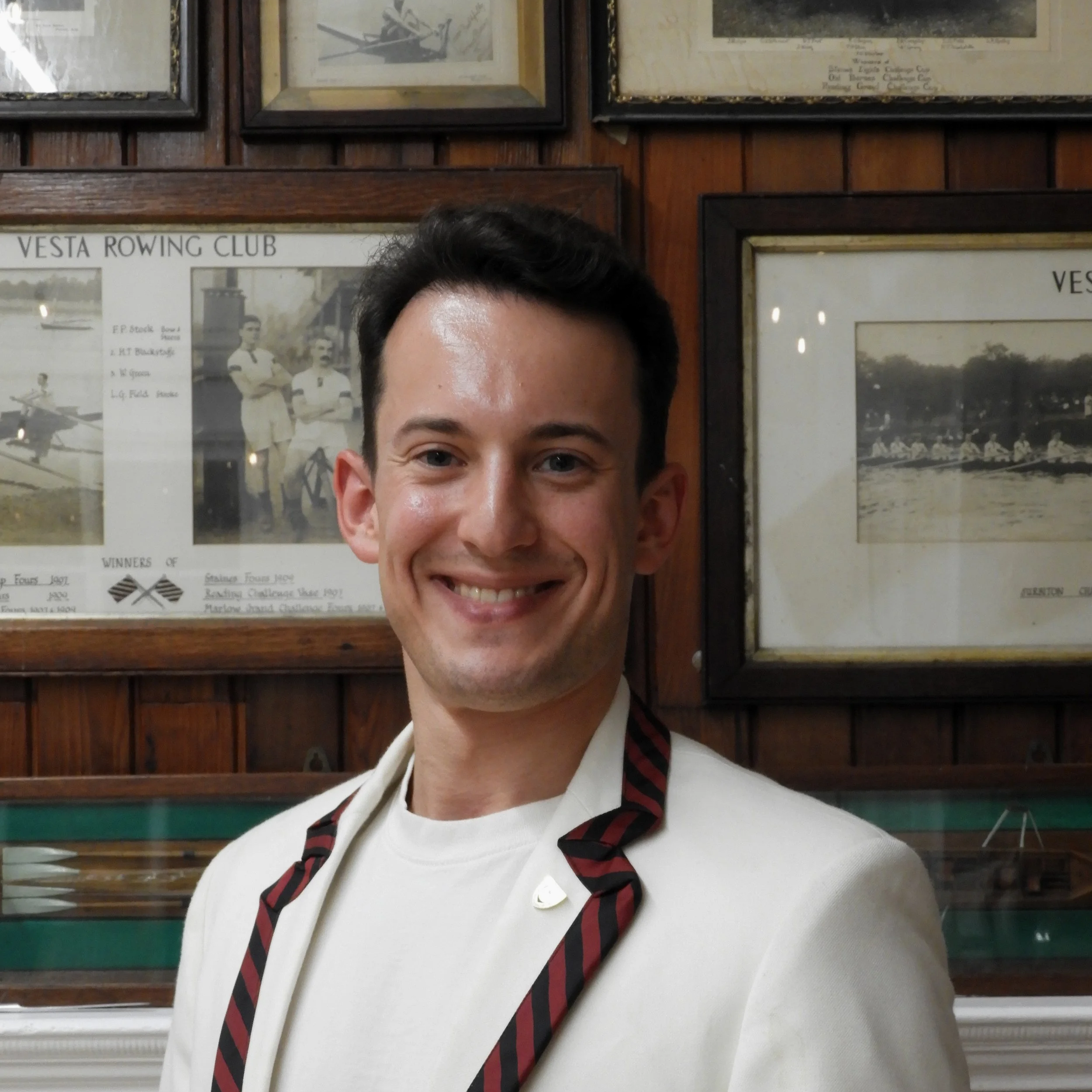Young man wearing a white shirt and black and red striped tie, smiling indoors with framed vintage photos on wooden-paneled walls behind him.