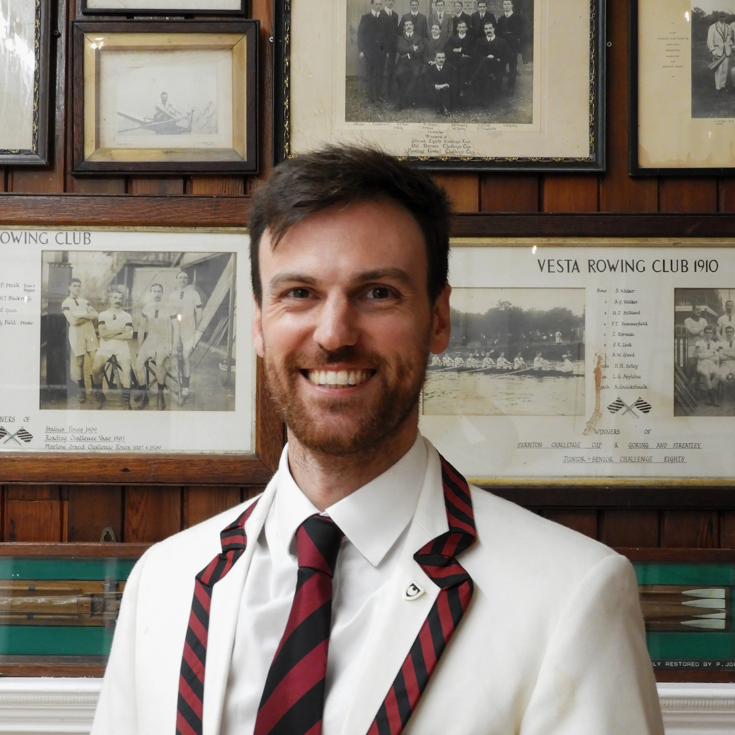 A man in a white shirt and tie standing in front of a wooden wall with framed black-and-white photos and posters related to rowing clubs from the early 1900s. His face is smiling, and only the upper part of his head and face are visible.