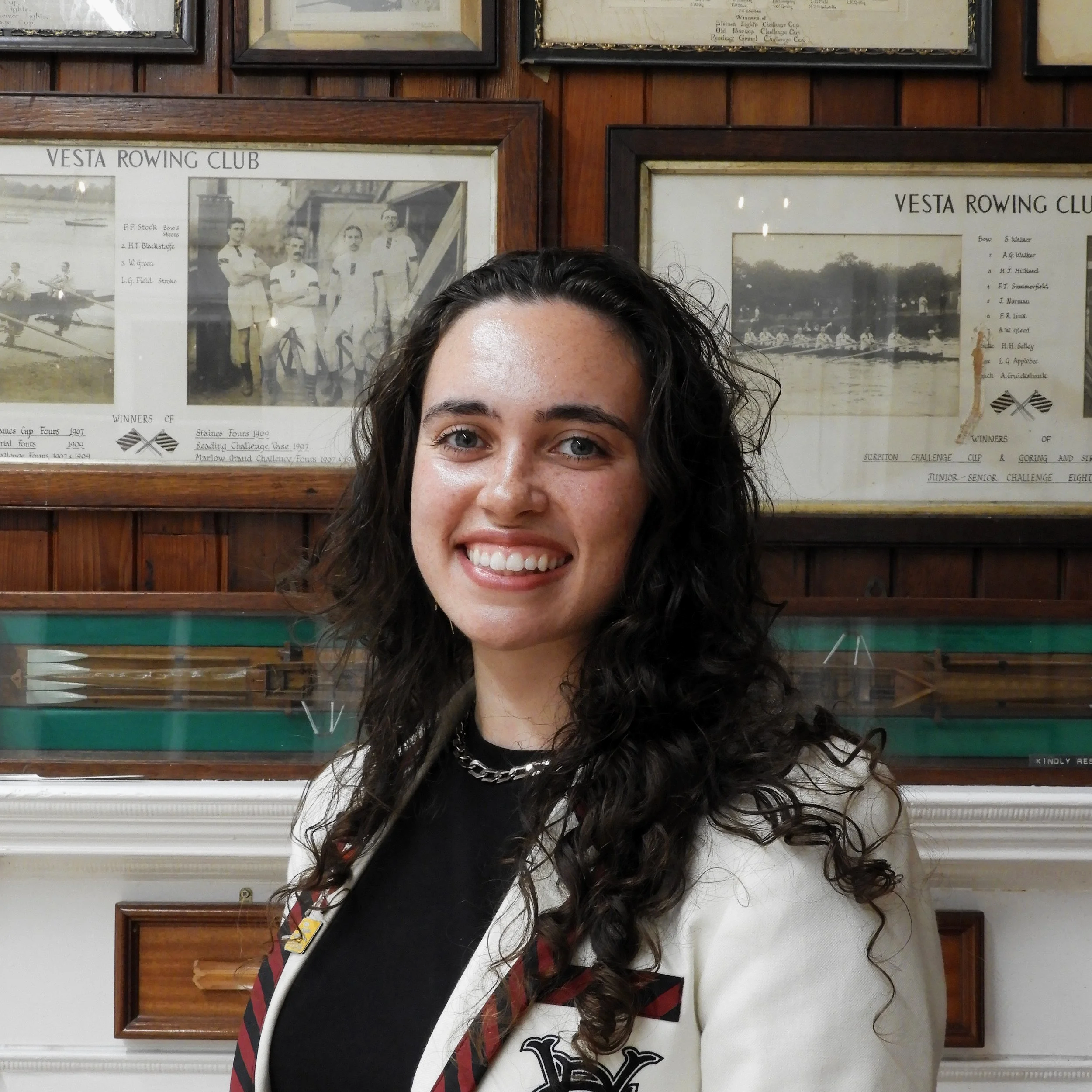 A woman with long curly dark hair smiling, standing in front of framed vintage photographs and newspaper clippings related to the Vesta Rowing Club on a wooden-paneled wall.