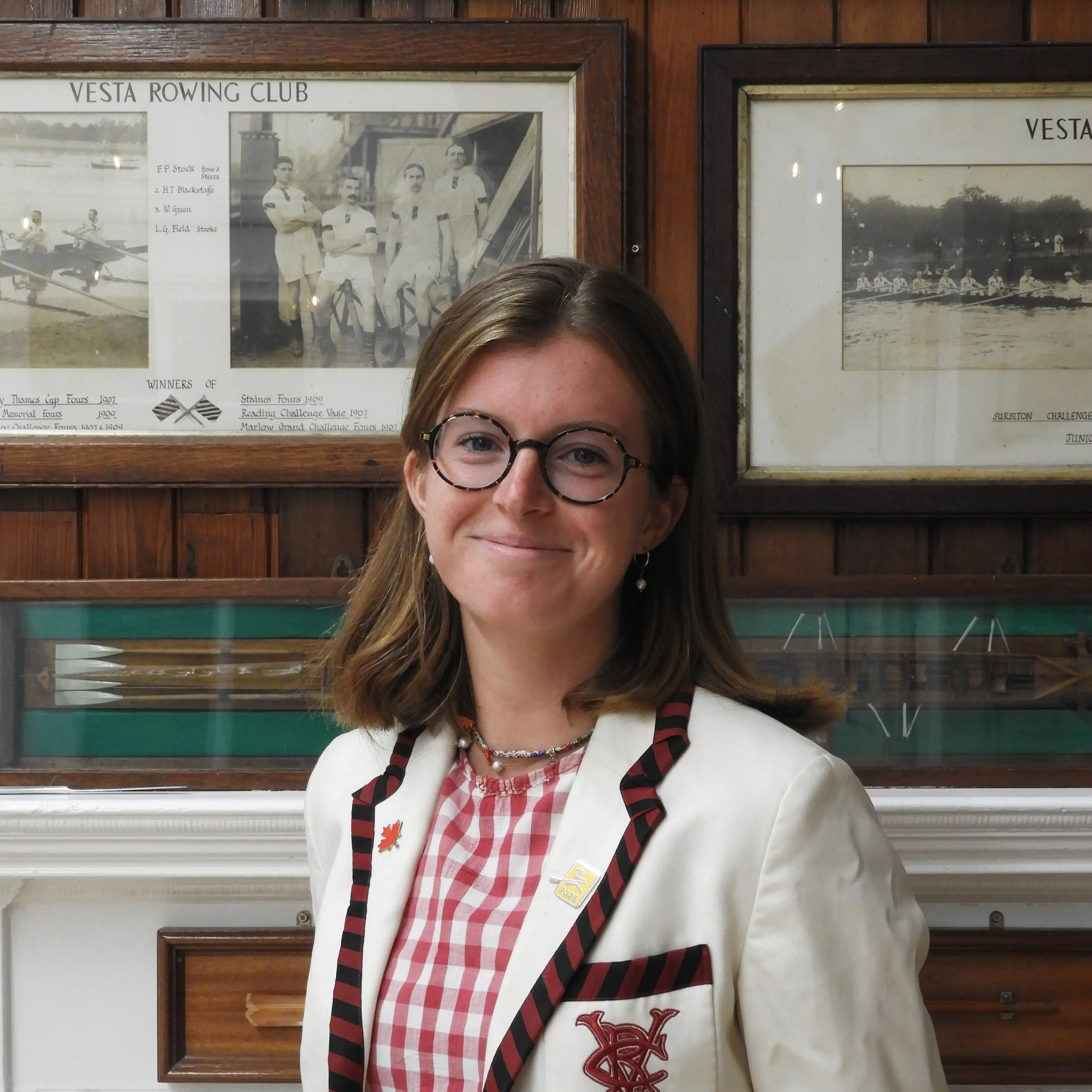 Close-up of a smiling woman with glasses wearing a white jacket with embroidered logos, standing in front of framed historical photographs related to Vesta Rowing Club on a wooden wall.