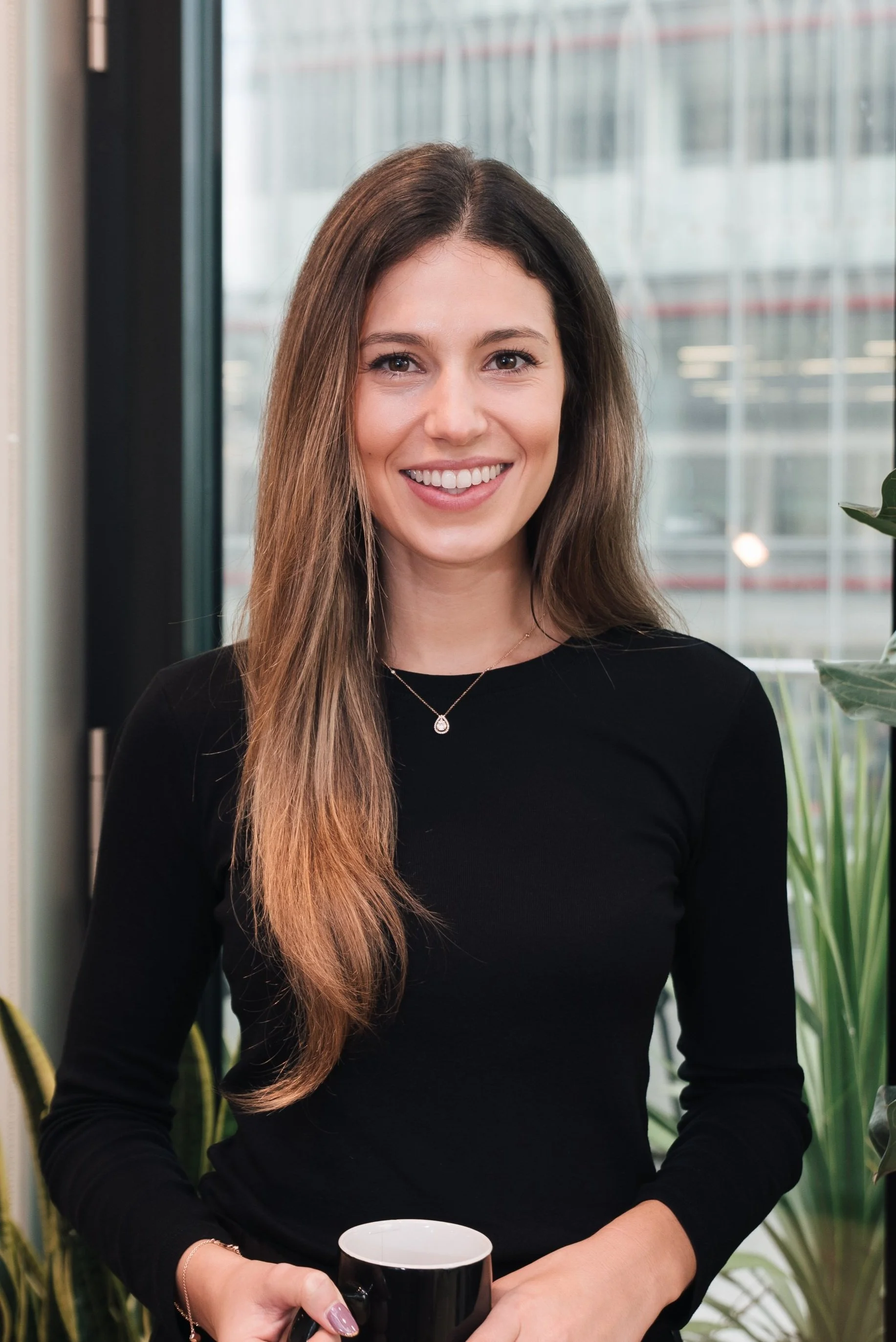 A young woman with long brown hair, smiling, holding a black coffee mug in a modern office or lounge setting, with large windows and green plants in the background.