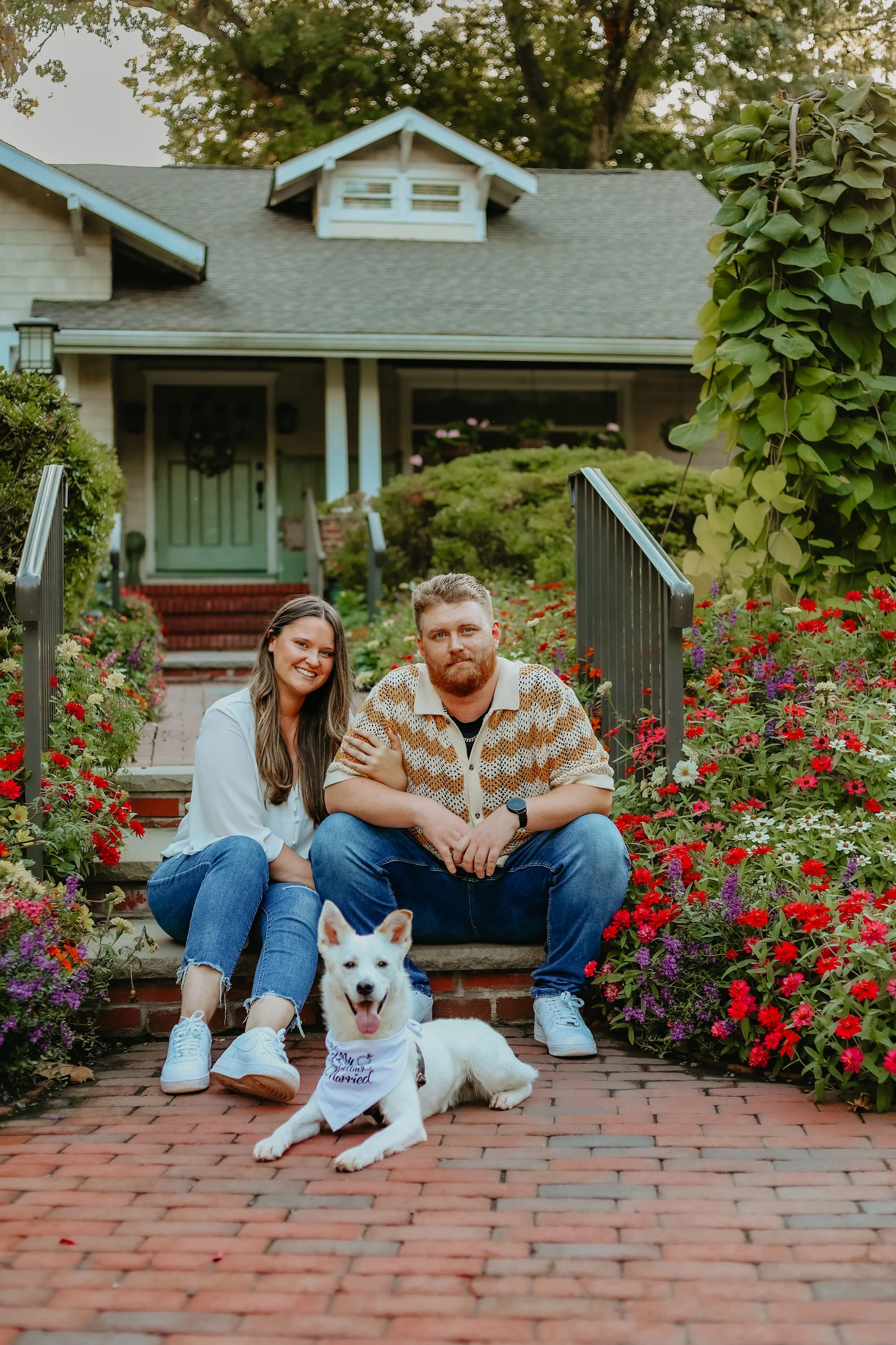 A happy couple with a white dog sitting on a brick pathway in front of their house, surrounded by colorful flowers and greenery.