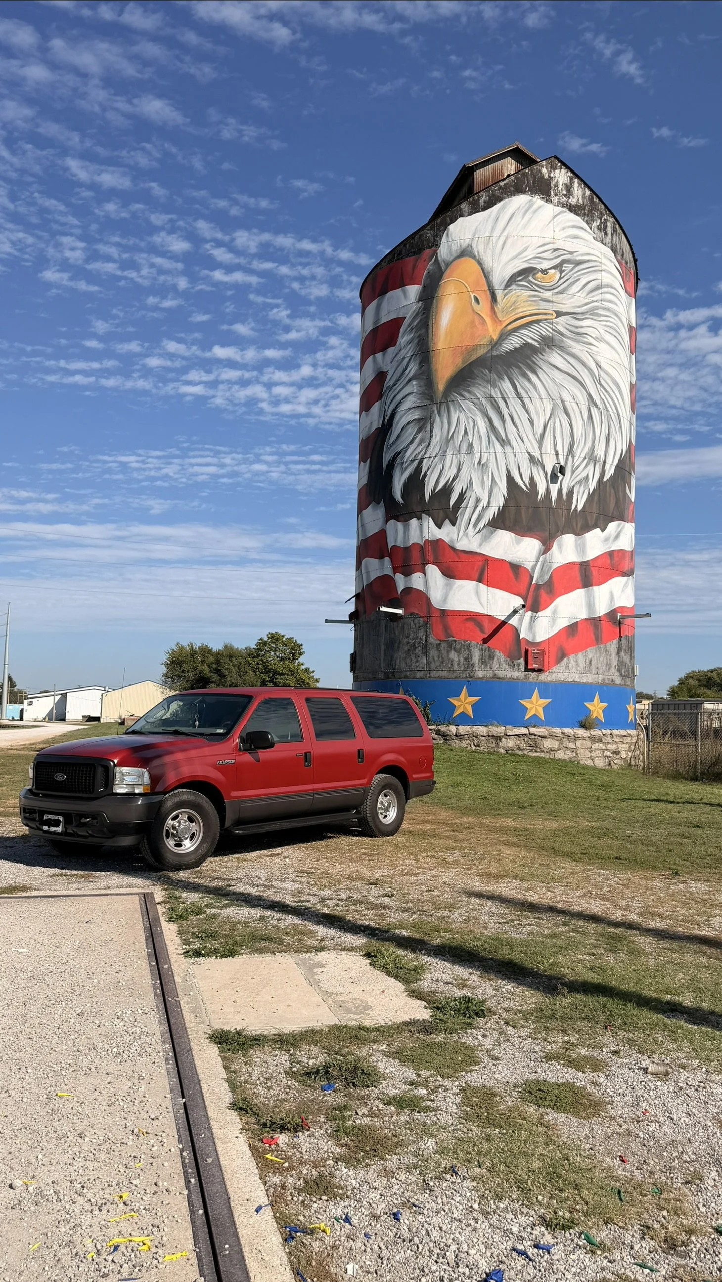 A large silo painted with a mural of a bald eagle wearing a red and white striped shirt, with a blue band and yellow stars at the bottom. A red Ford excursion vehicle is parked in front of the silo on a grassy area.