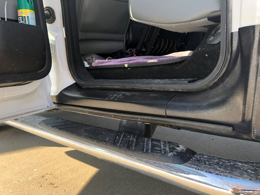 View of the interior of a pickup truck from the passenger side, showing the step, black interior trim, and part of the back seat with a pink towel and some items. The door is open, revealing the truck's black interior and a dusty step.